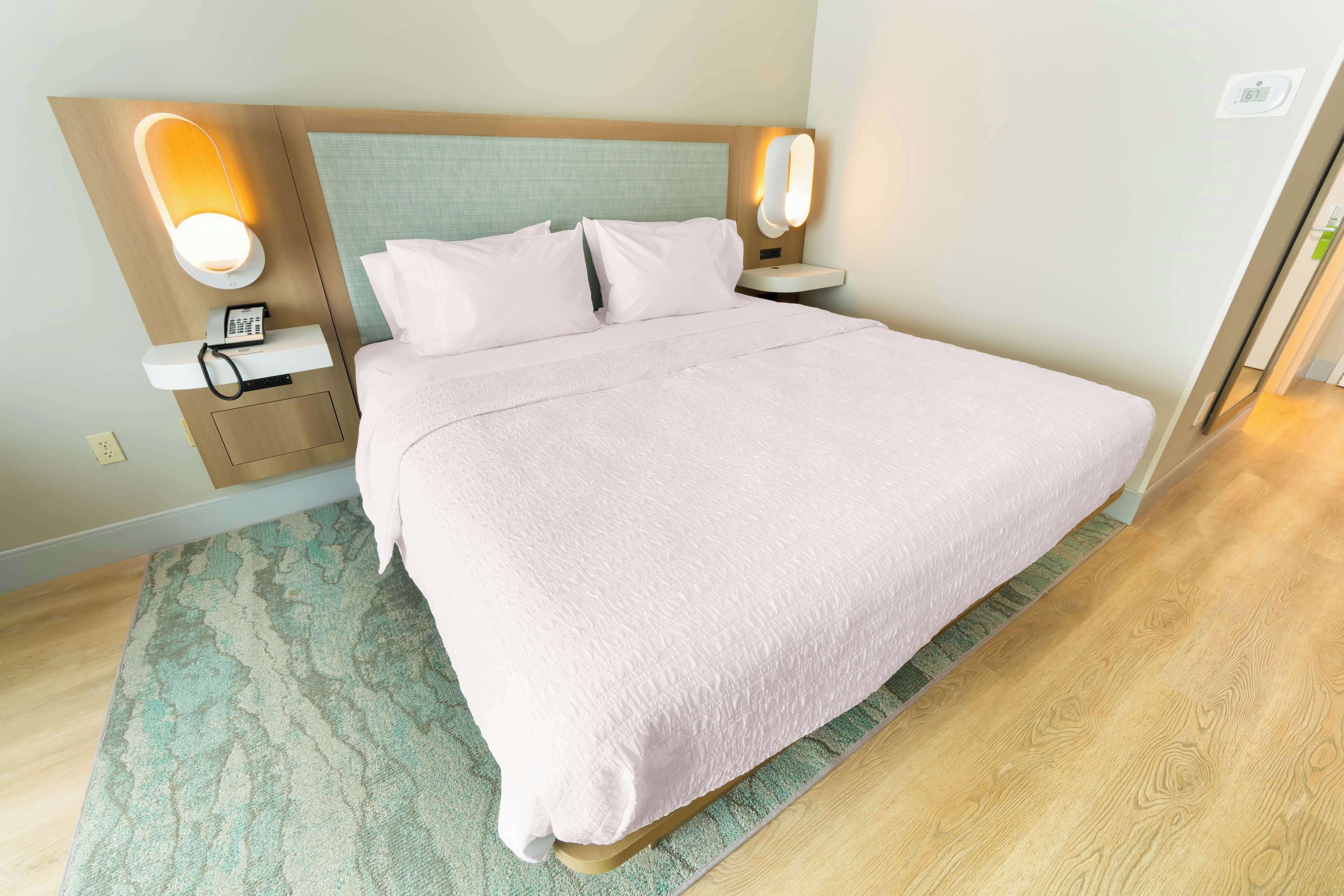 Een netjes opgemaakt bed met een lichtgekleurde deken en kussens, een nachtkastje met een lamp, en een aan de muur bevestigde telefoon.