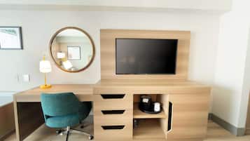 A modern hotel room with a wooden desk, a teal office chair, a round mirror, and a mounted flat-screen TV.