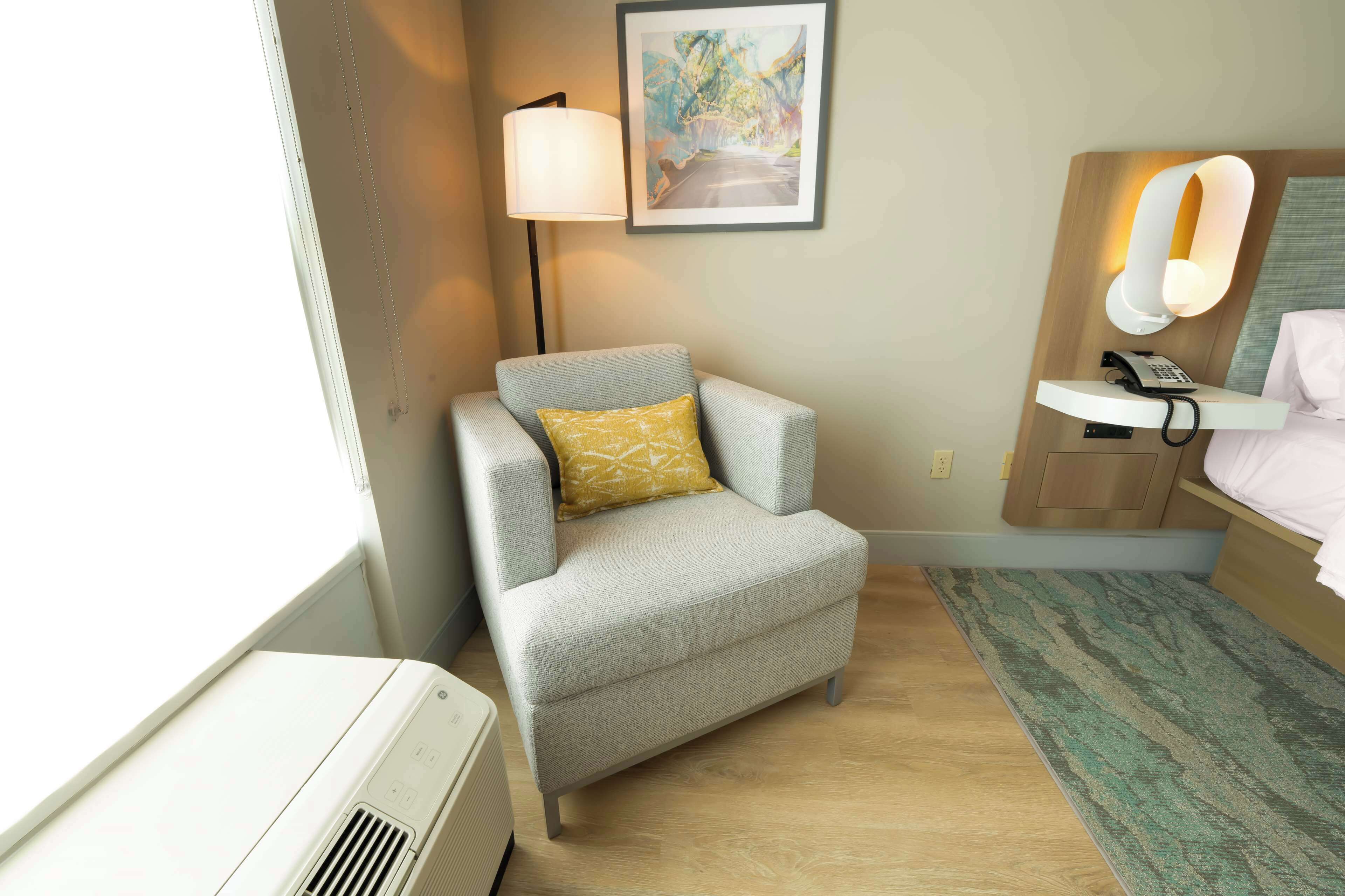 A hotel room with a grey armchair, a bedside table with a lamp, a phone, and a bed with a headboard.
