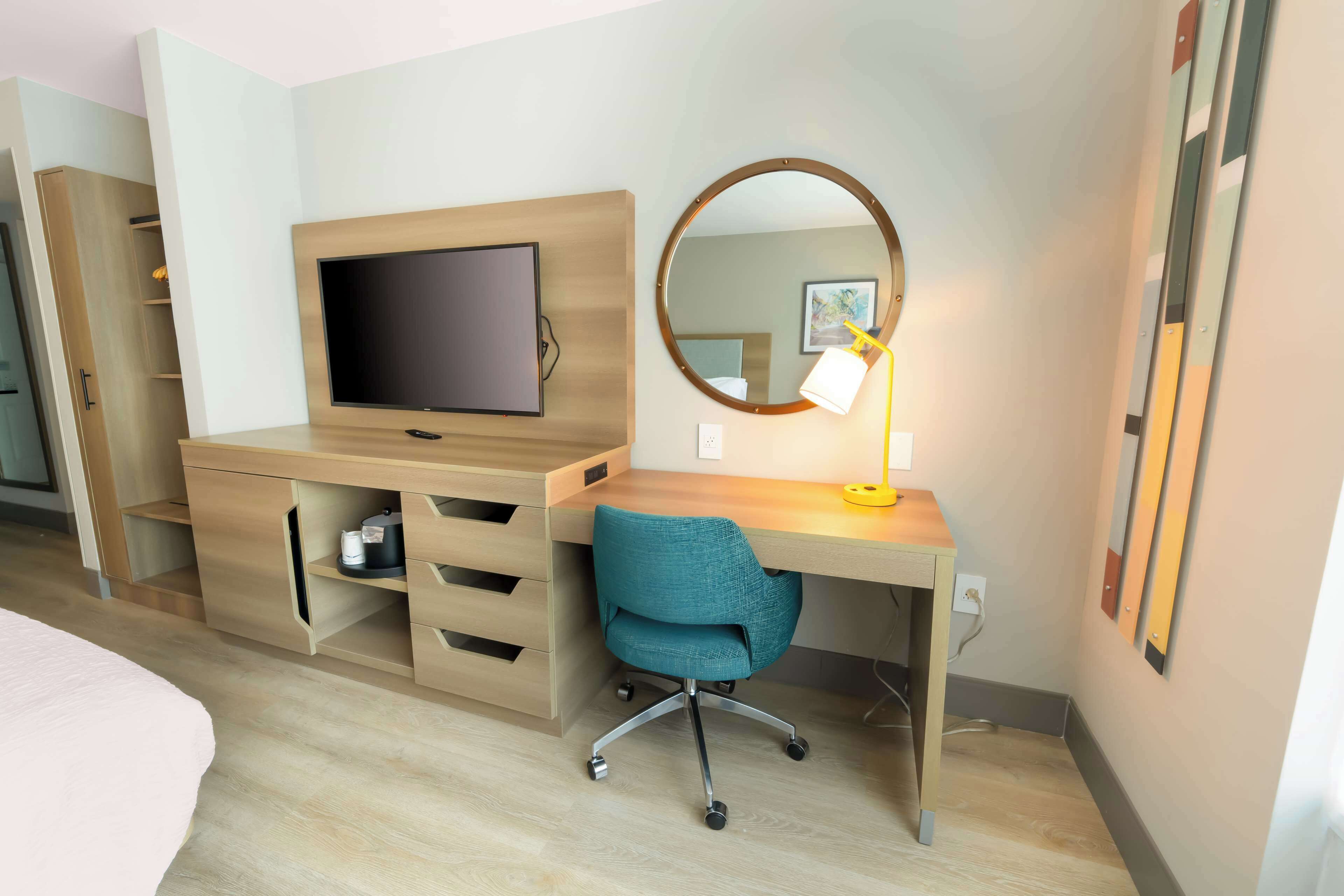 A modern hotel room with a wooden desk, a television, a round mirror, and a teal office chair.