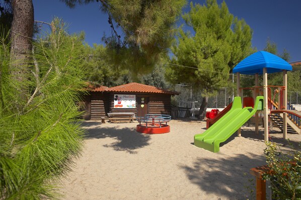 A playground with a wooden playhouse, a red and green slide, and a sandy ground. - Portes Beach Hotel (Nea Propontida)