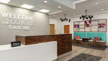 A hotel reception area with a 'Welcome to the Coyote State' sign, a wooden reception desk, a teal sofa, and a black rug.