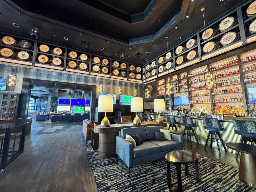 Zona de bar con diversas opciones de asientos, exhibición de botellas de licor y una pared de barriles de madera.