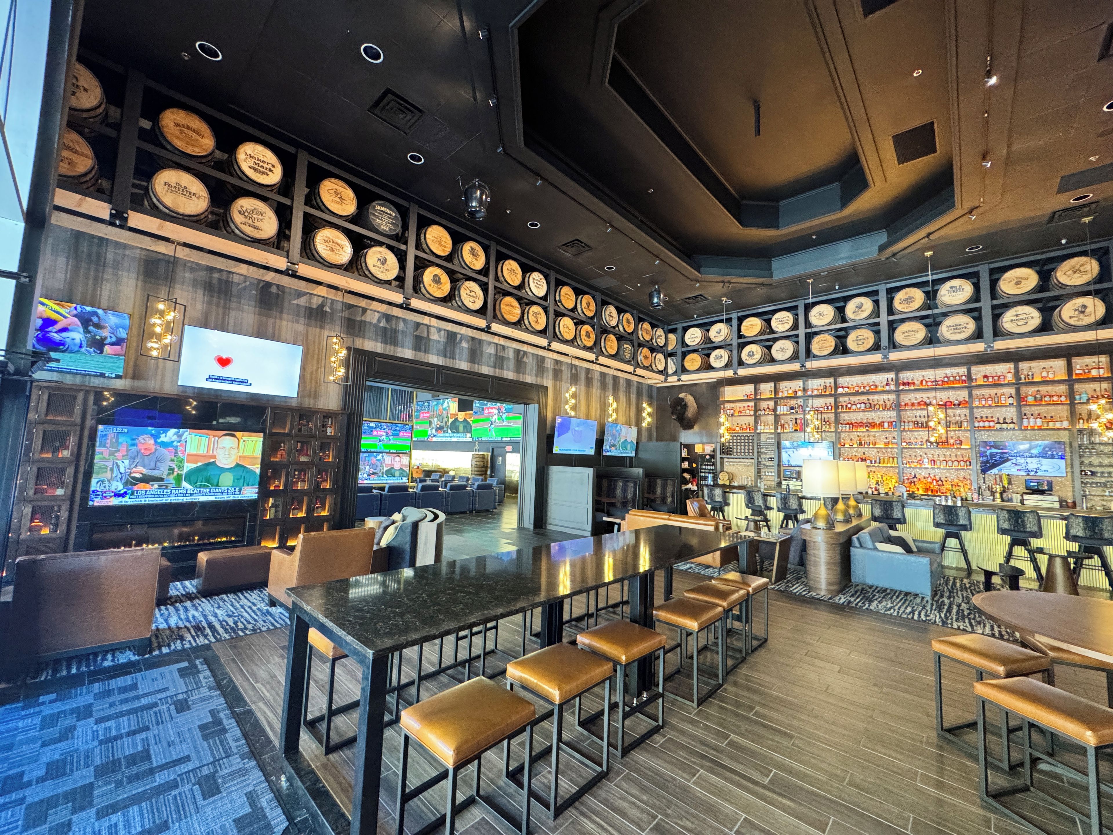 A bar area with multiple seating options, a television screen displaying sports, and a wall adorned with numerous bottles.