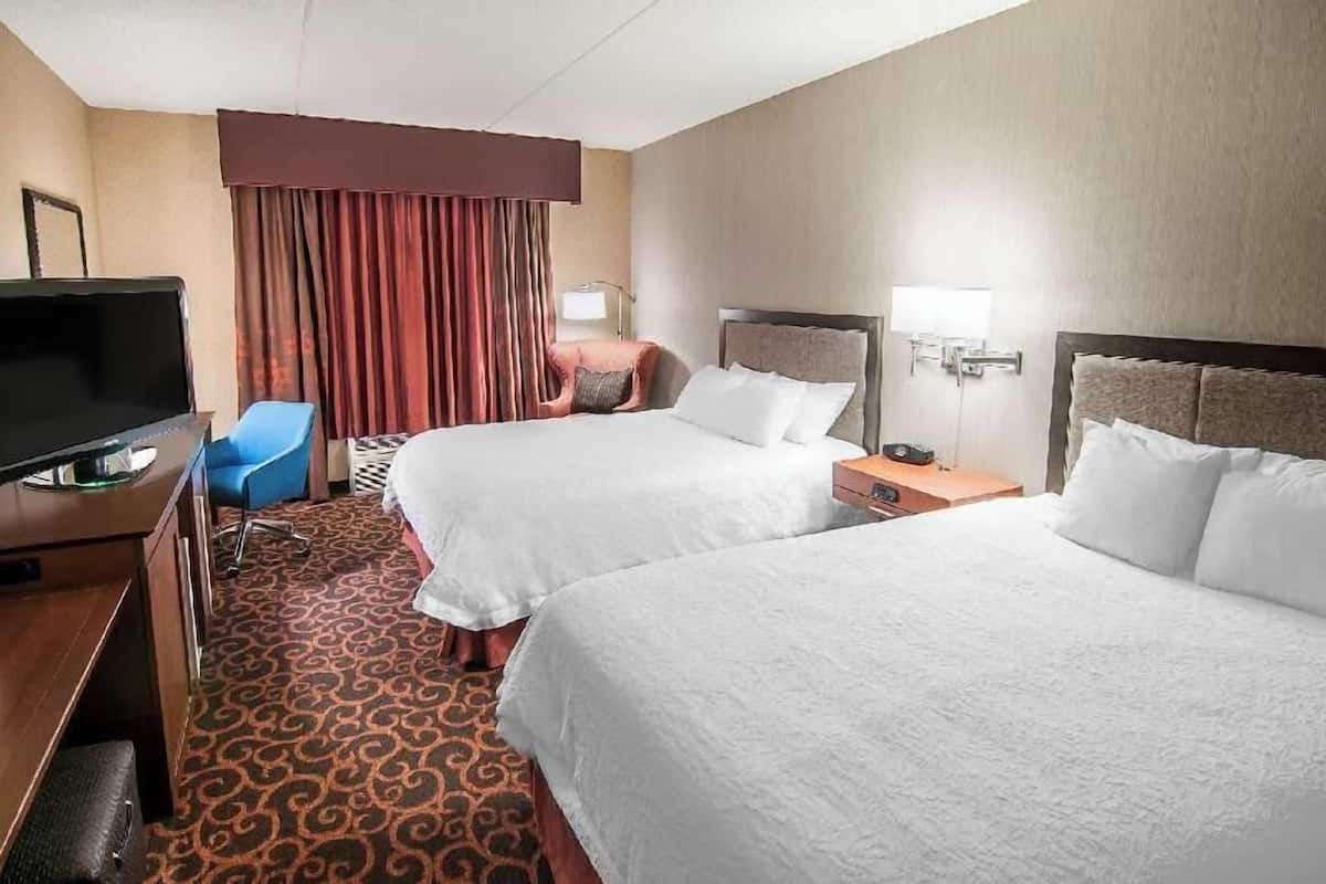 Habitación de hotel con dos camas, un escritorio con televisor, una silla, una lámpara de escritorio y una ventana con cortinas.