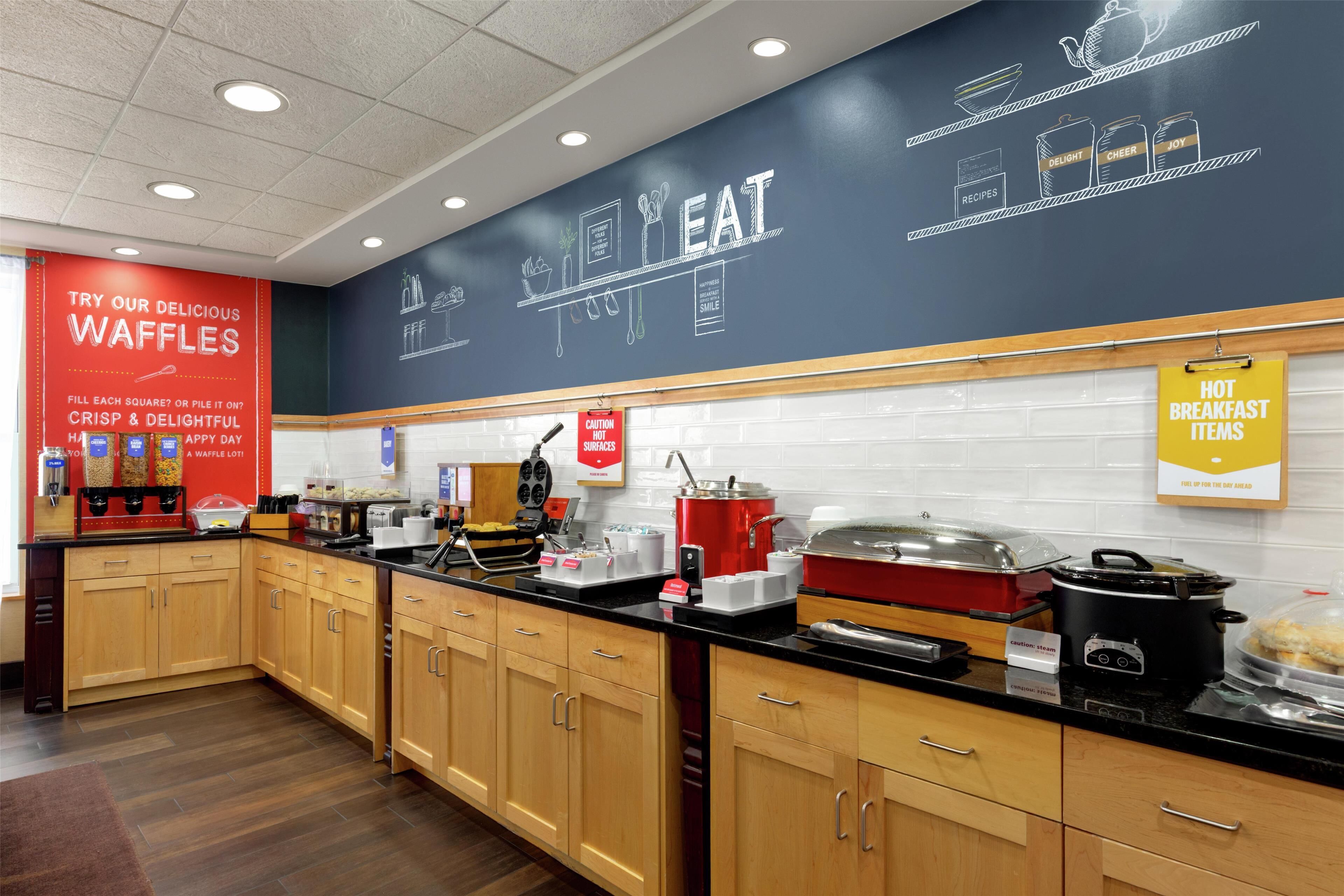 A kitchen area with wooden cabinets, a black countertop, and various appliances like a coffee maker and a slow cooker. There are signs for waffles and hot breakfast items.