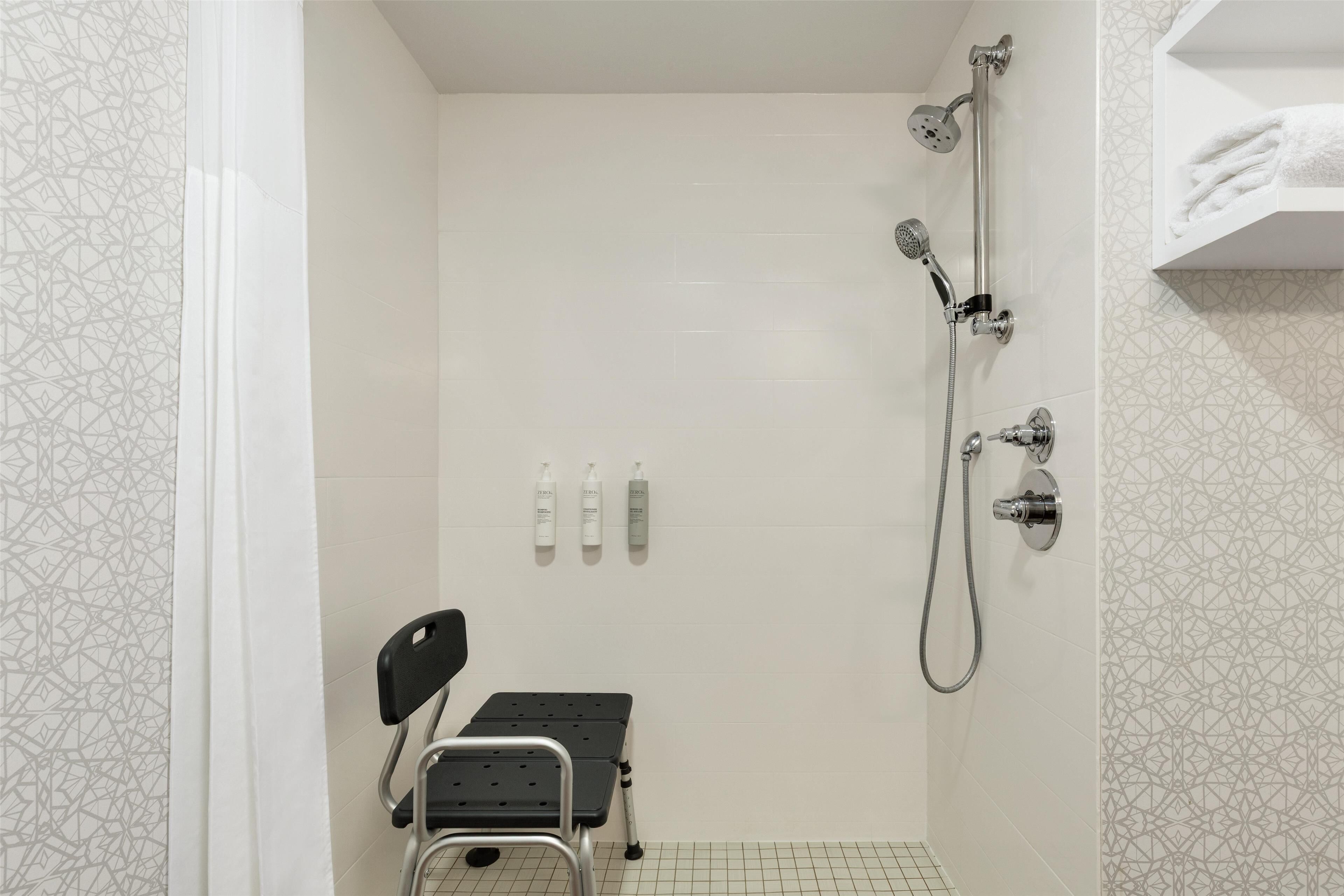 Un espacio de ducha con cabezal de ducha de mano, grifo empotrado en la pared y otro cabezal de ducha de mano. Hay un estante pequeño con una toalla y una botella de champú.