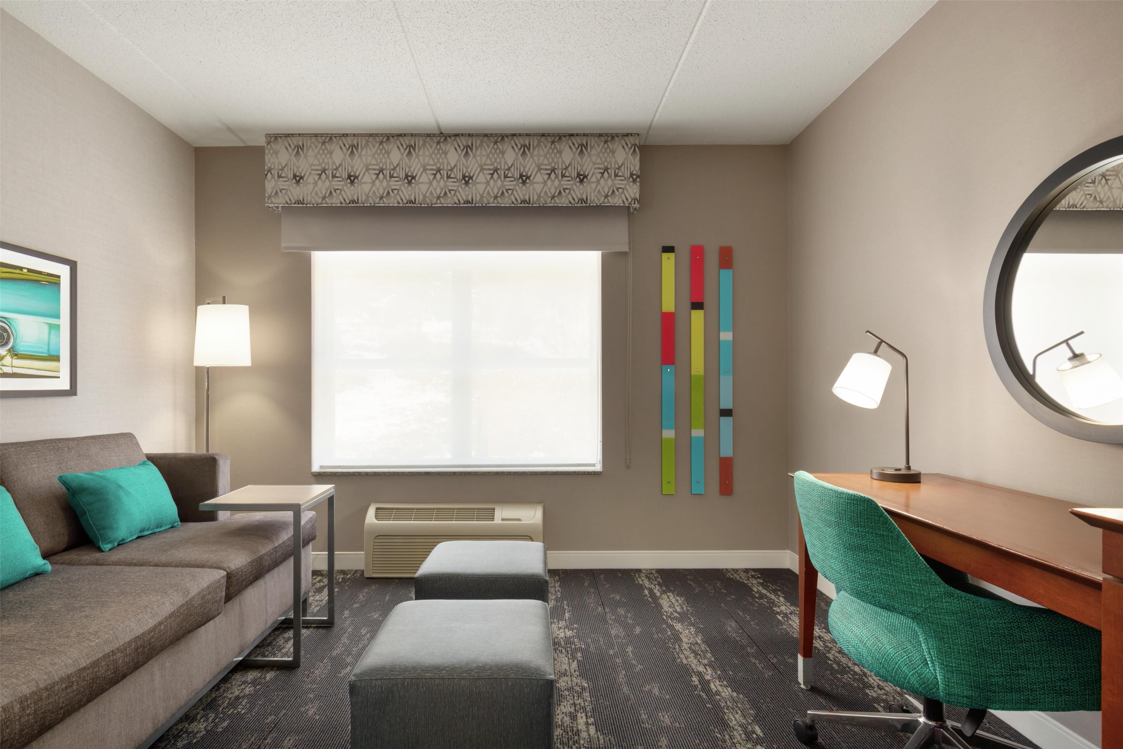 A hotel room with a sofa, ottoman, desk, chair, lamp, air conditioning unit, window, and a wall with colorful decor.