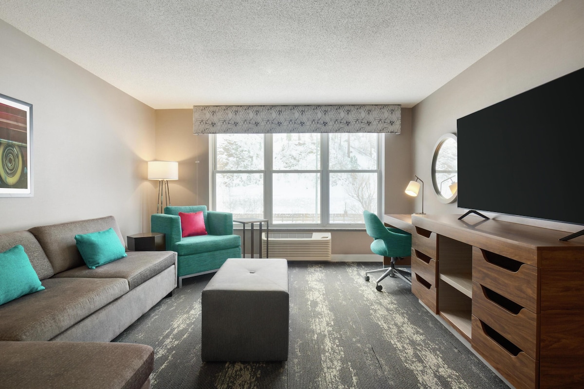 A modern hotel room with a grey sofa, teal armchairs, a wooden desk with a flat-screen TV, and a large window with a view of trees.