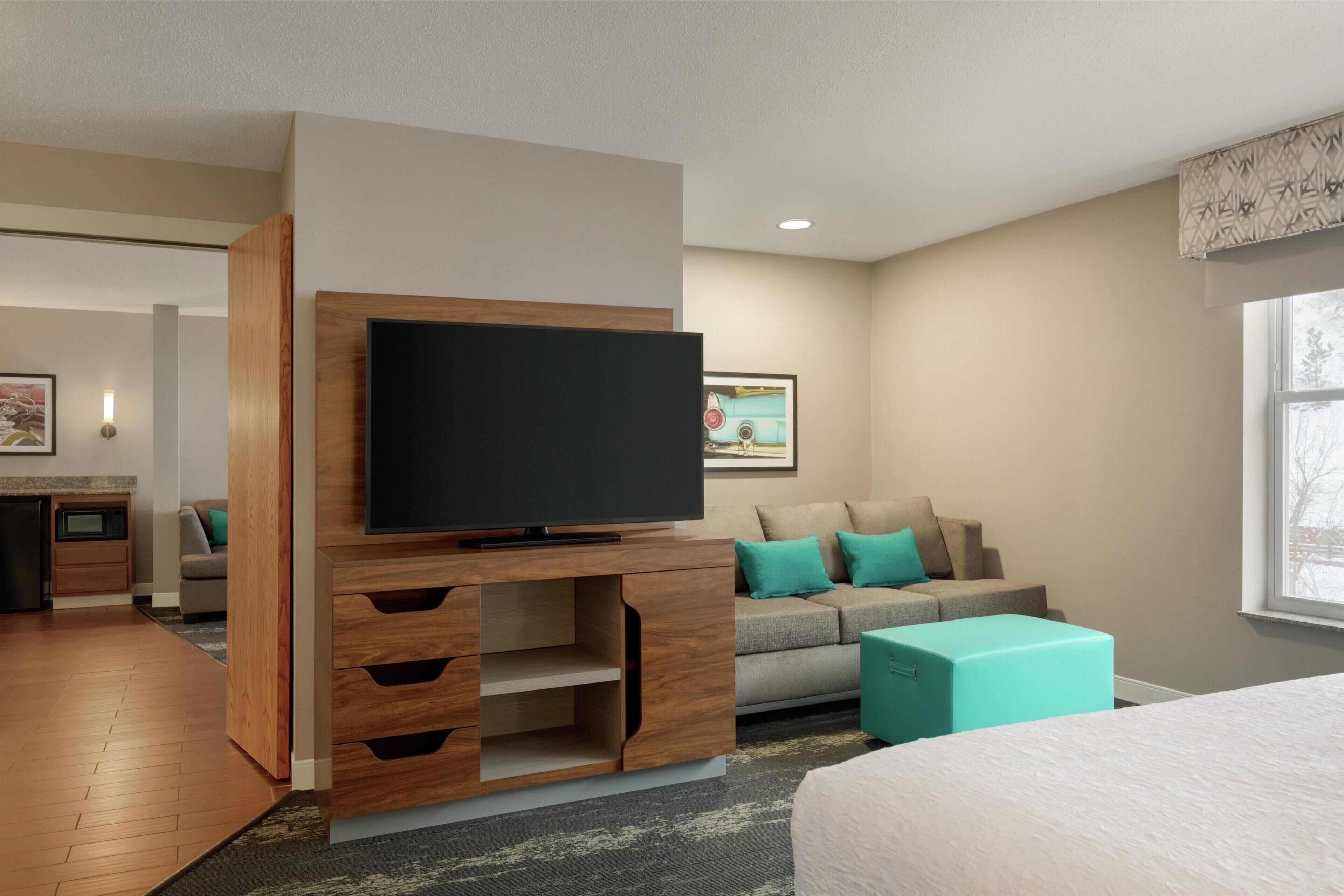 Une chambre d’hôtel moderne dotée d’un téléviseur à écran plat installé sur un meuble bas en bois, d’un canapé avec des coussins turquoise, d’un lit et d’une fenêtre offrant une vue agréable.