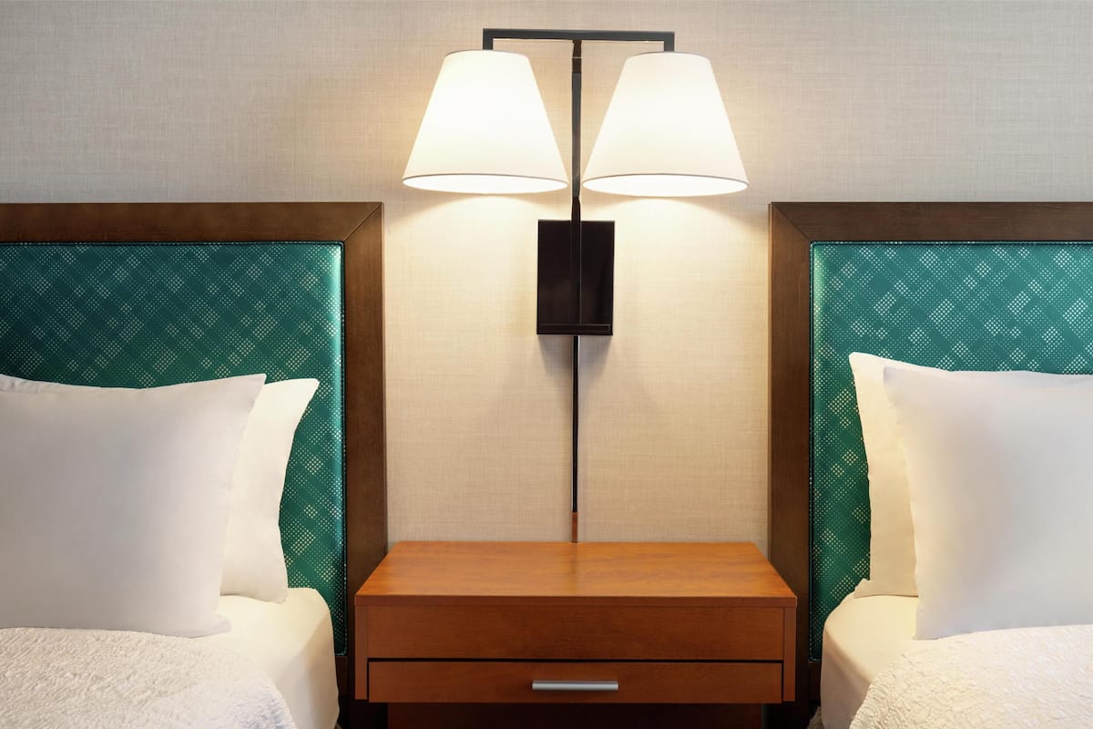 Habitación de hotel con dos camas, cada una con cabecero verde, mesita de noche de madera con cajón y una lámpara de pared entre las camas.