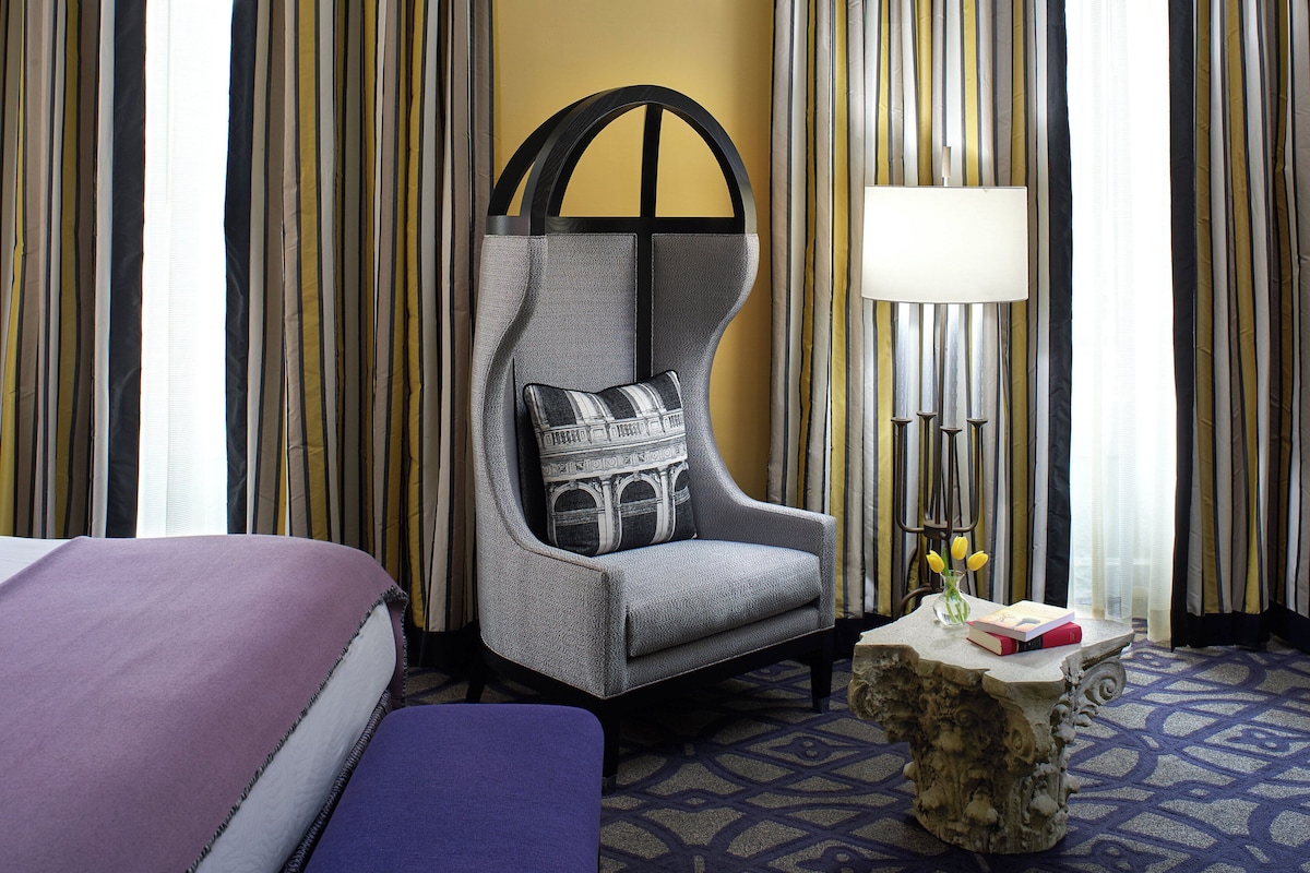 A hotel room with a bed, a chair with a patterned cushion, a small table with a vase of flowers, and striped curtains.