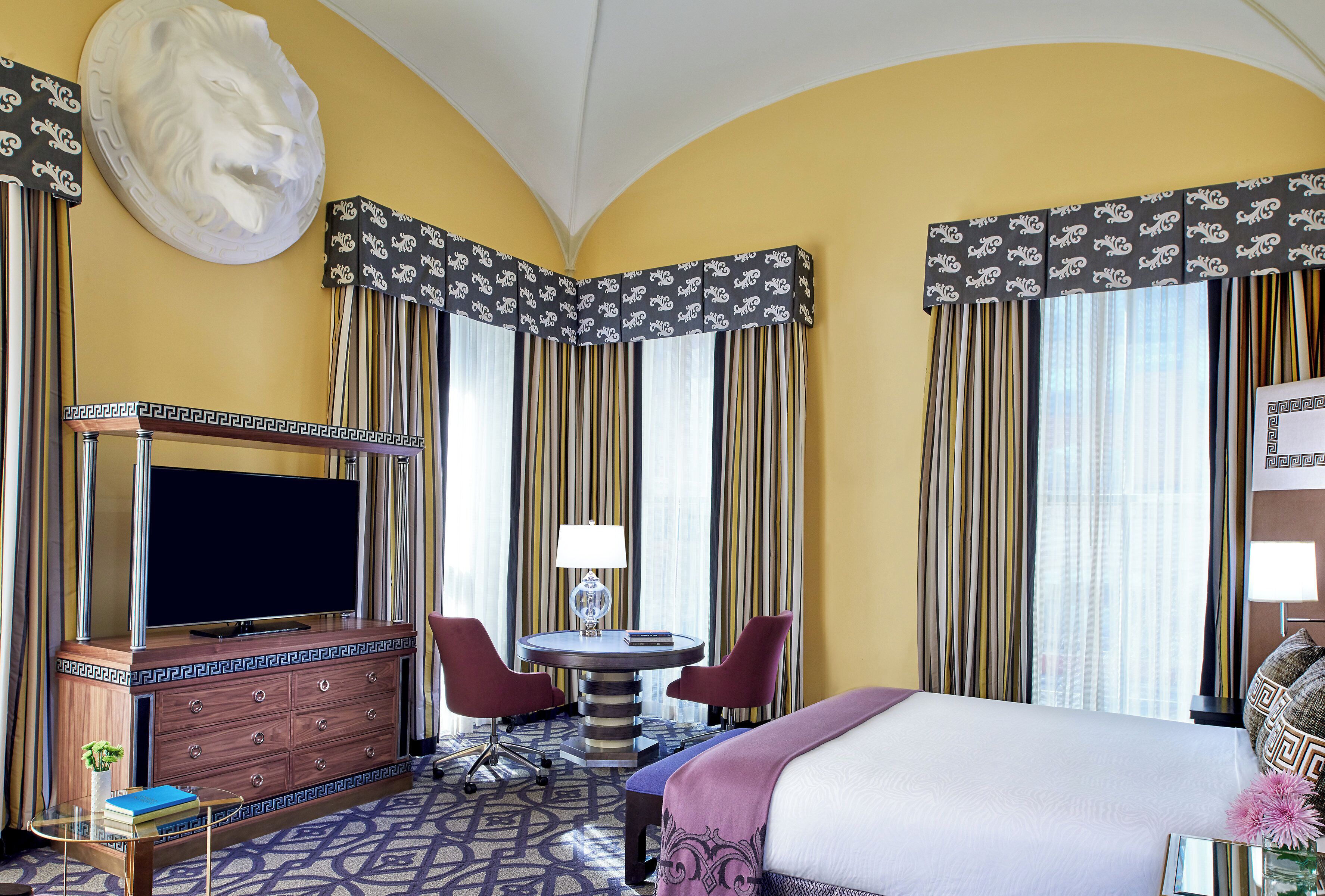 A hotel room with a bed, a TV, a desk with a lamp, a chair, and a decorative headpiece on the wall.