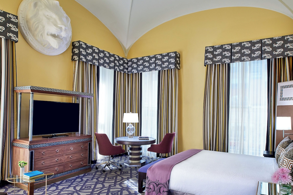A hotel room with a bed, a TV, a desk with two chairs, a dresser, and curtains.
