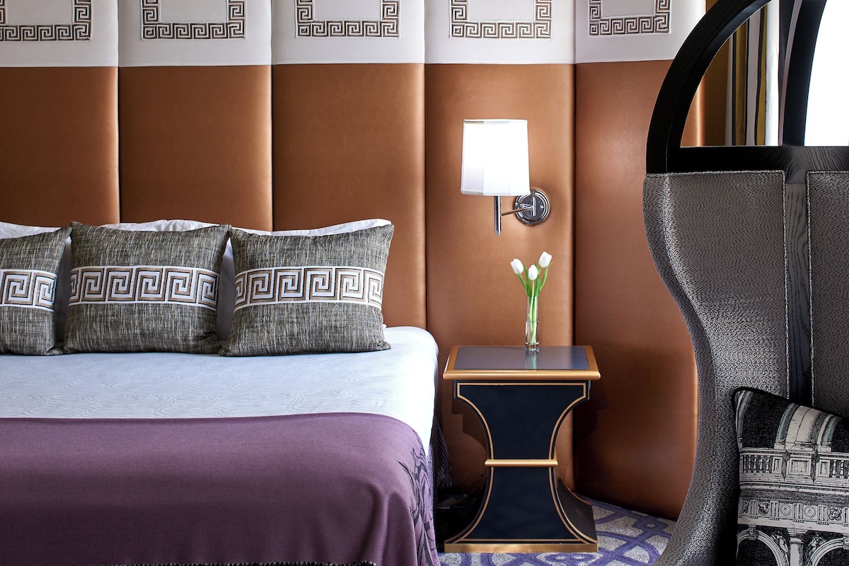 A hotel room with a bed, a nightstand, a lamp, a vase with flowers, and a chair.