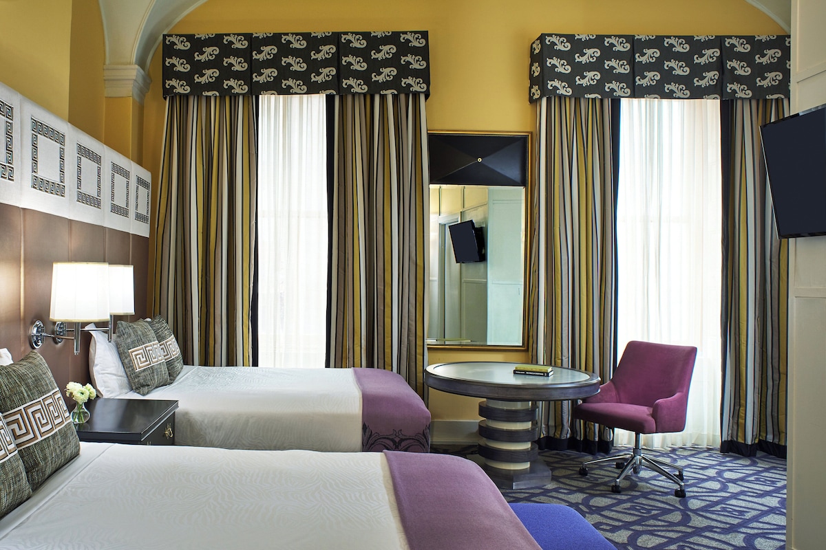 A hotel room with two beds, a round table, a chair, a TV, and a mirror.