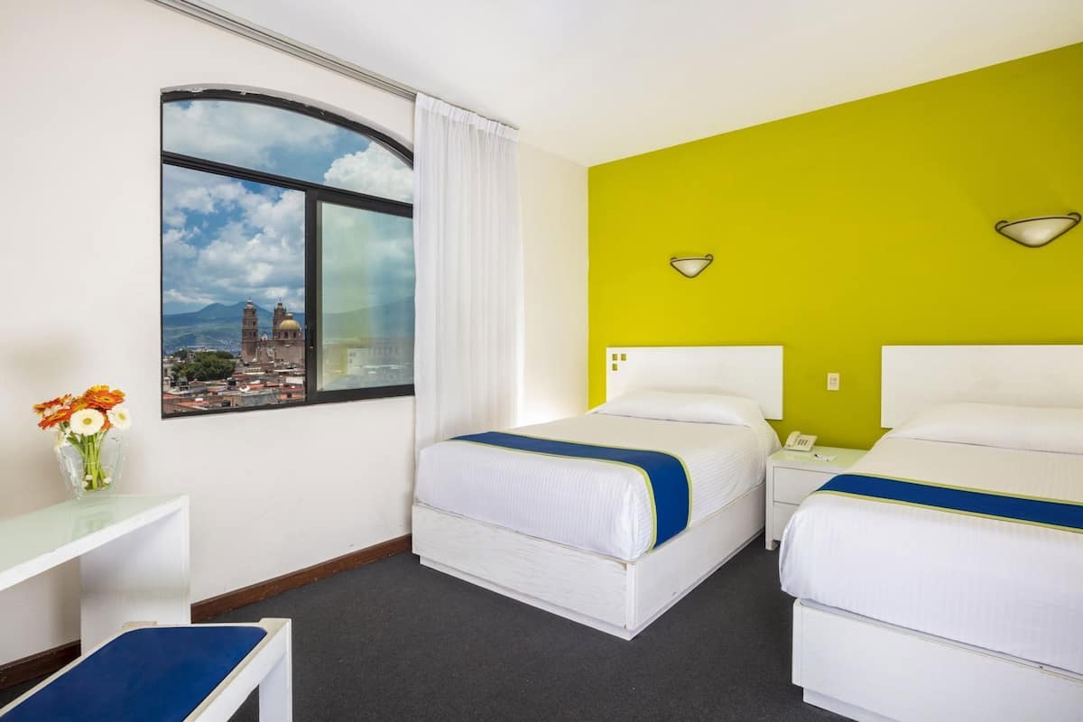 A hotel room with two beds, a window with a cityscape view, a desk with a vase of flowers, and a bright green accent wall.