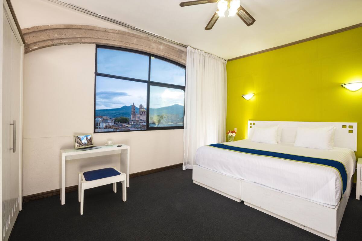 A hotel room with a large window, a bed, a bedside table, and a desk.