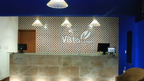 A reception area with a patterned wall, a reception desk, and a wicker chair. - Hotel Vista Express Morelia By Arriva Hospitality Group (Morelia)