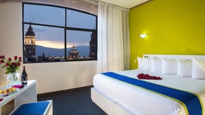 A hotel room with a large bed, a desk with a vase of flowers, and a view of a cityscape through a large window. - Hotel Vista Express Morelia By Arriva Hospitality Group (Morelia)