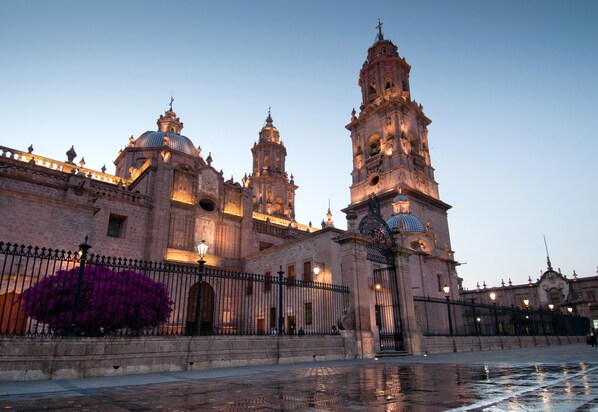 A historic building with multiple towers and domes, a gated entrance, and a reflective water surface in front.