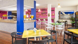 A brightly colored restaurant with a central counter, multiple tables and chairs, and decorative wall patterns. - Hotel Vista Express Morelia By Arriva Hospitality Group (Morelia)