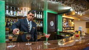 A bar area with a bartender pouring a drink, a variety of bottles on shelves, and several cocktails on the counter.
