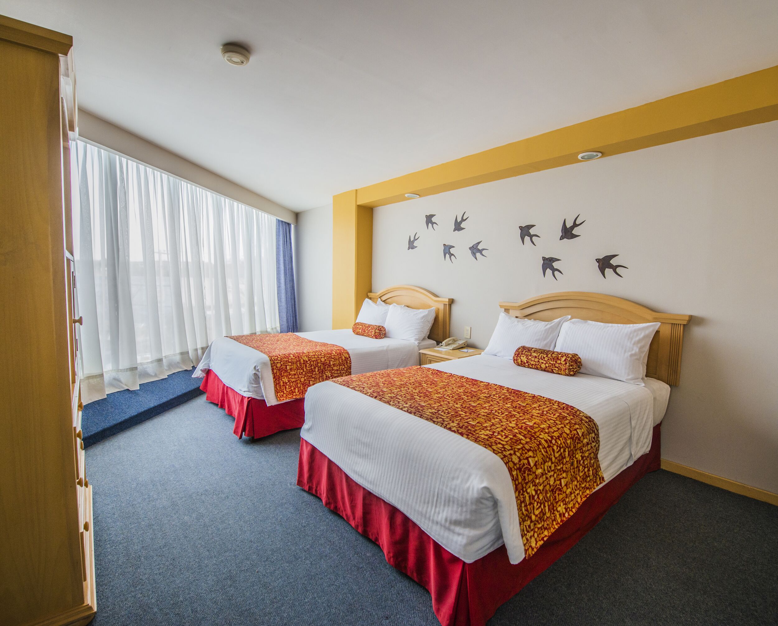 Habitación de hotel con dos camas, cabecera de madera, pared con siluetas de pájaros y ventana con cortinas translúcidas.