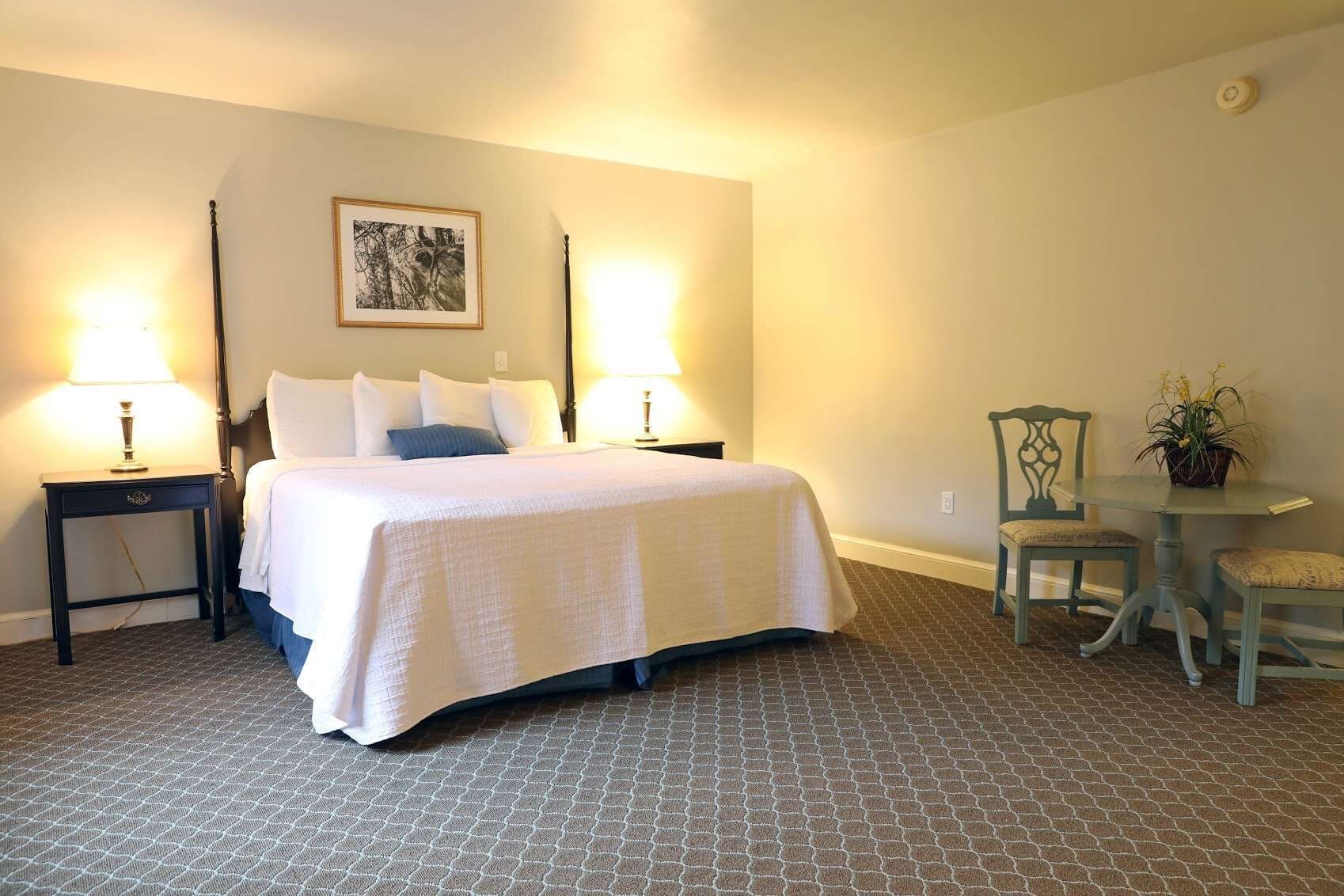 Habitación de hotel ordenada con cama, dos mesitas de noche con lámparas, una pequeña mesa con sillas y un cuadro en la pared.
