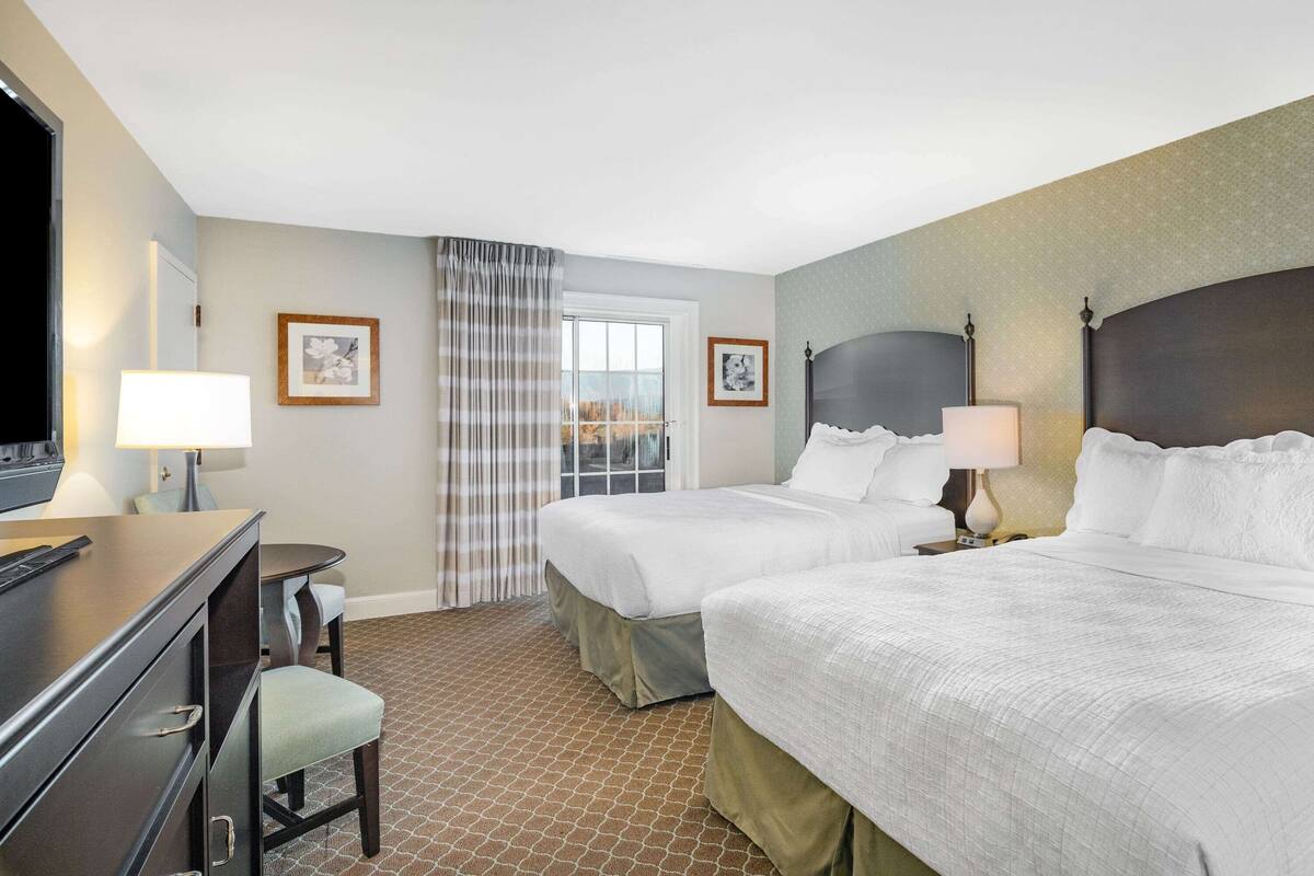 Habitación de hotel con dos camas, un escritorio, una silla y un ventanal con cortinas.