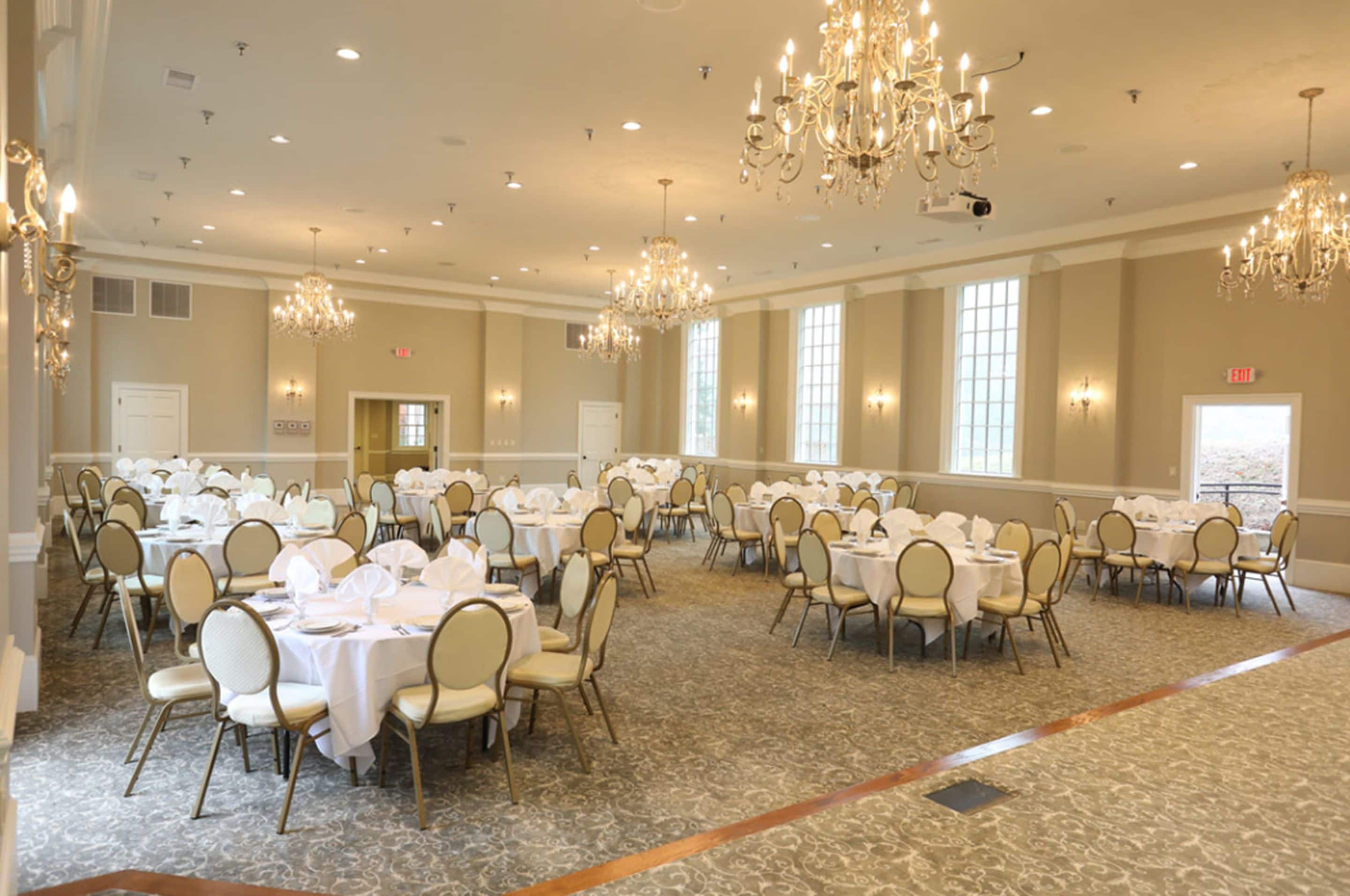 A banquet hall with round tables, chairs, chandeliers, and large windows.