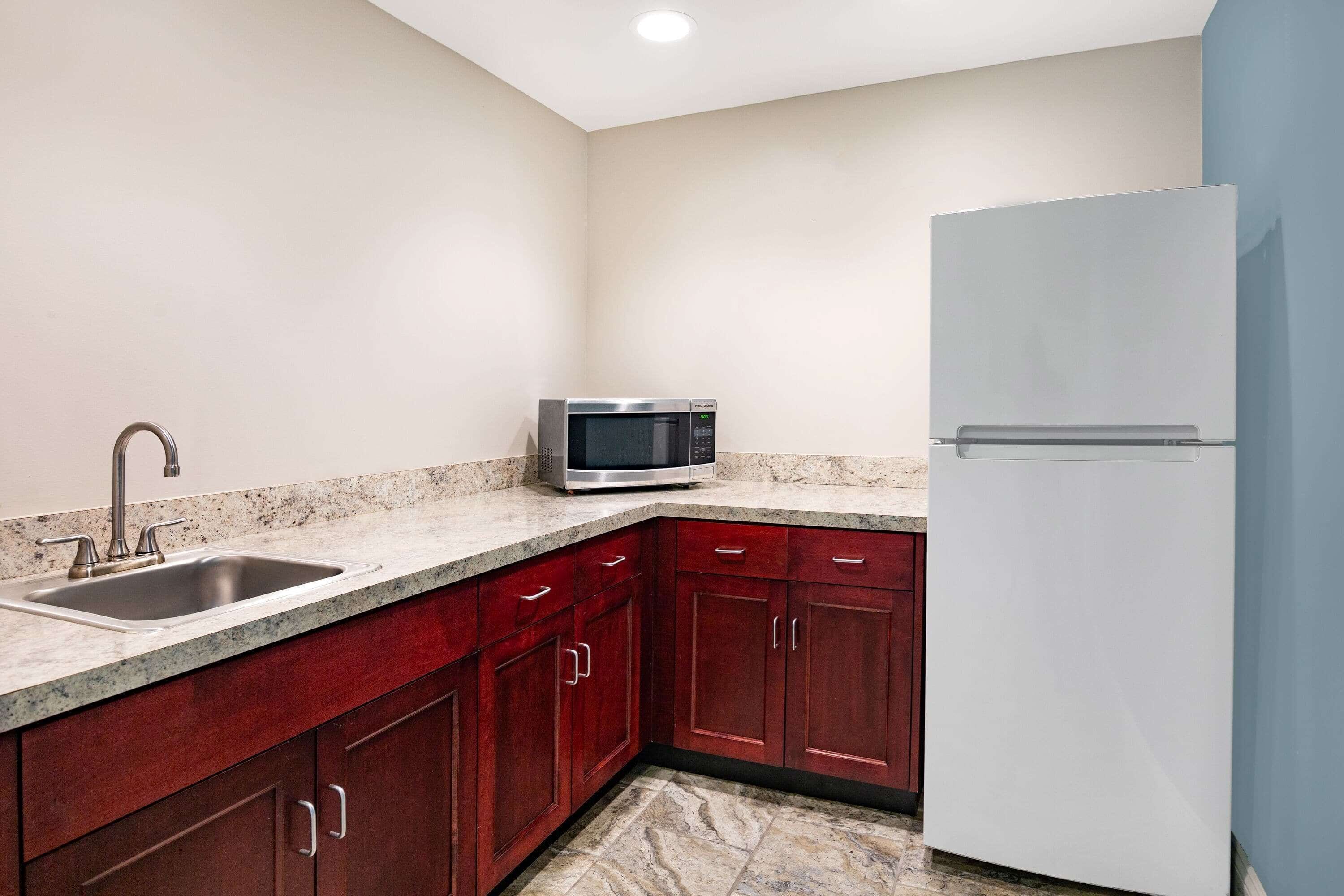 A kitchen with a granite countertop, stainless steel sink, red cabinets, a microwave, and a white refrigerator.