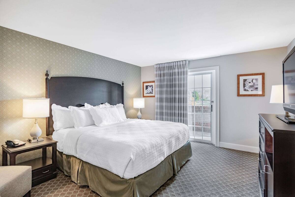 Una habitación de hotel con una cama grande, dos lámparas de noche, una mesita de noche, un aparador y una ventana con cortinas.
