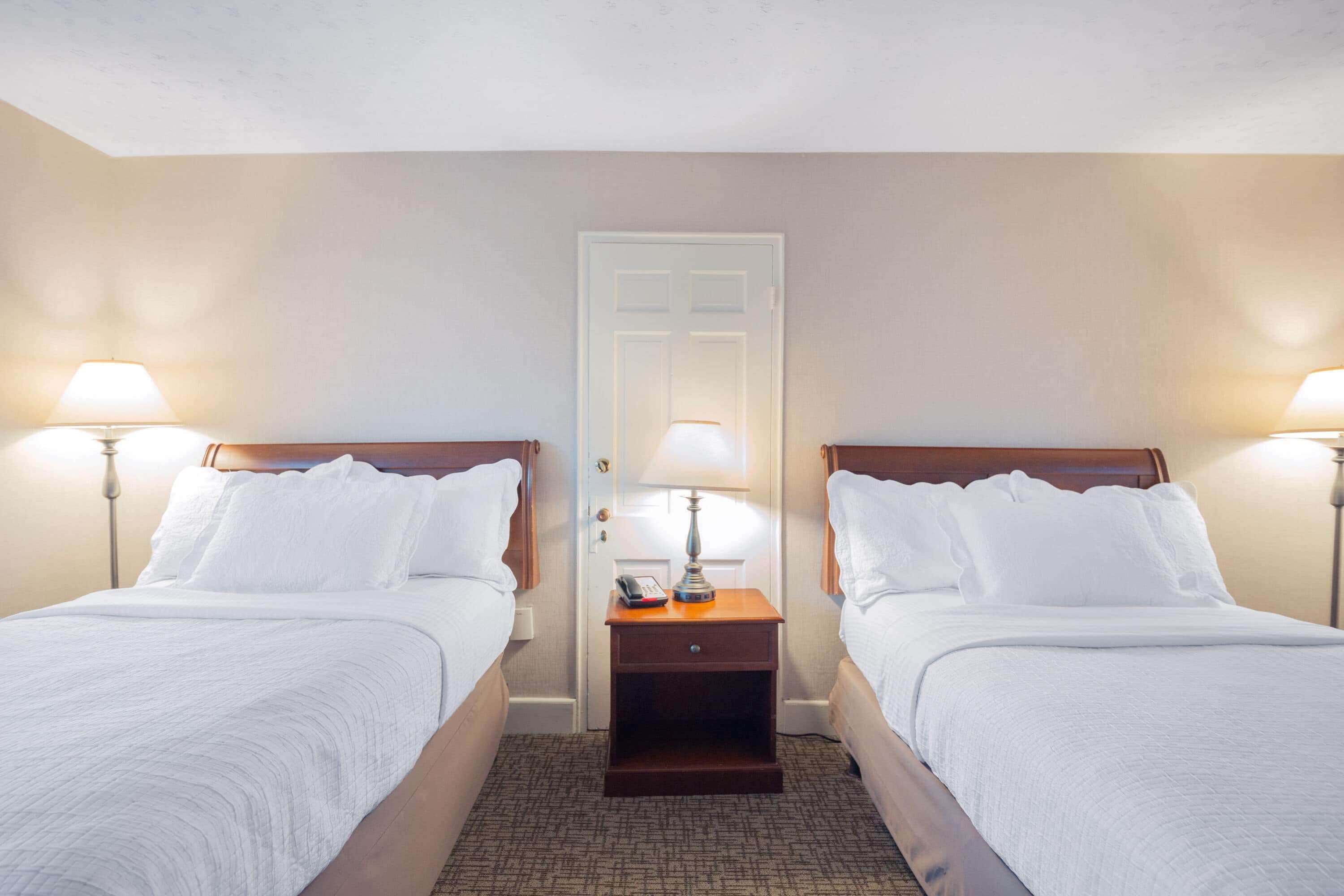A hotel room with two beds, each with white linens, a wooden headboard, and a nightstand with a lamp and a phone.