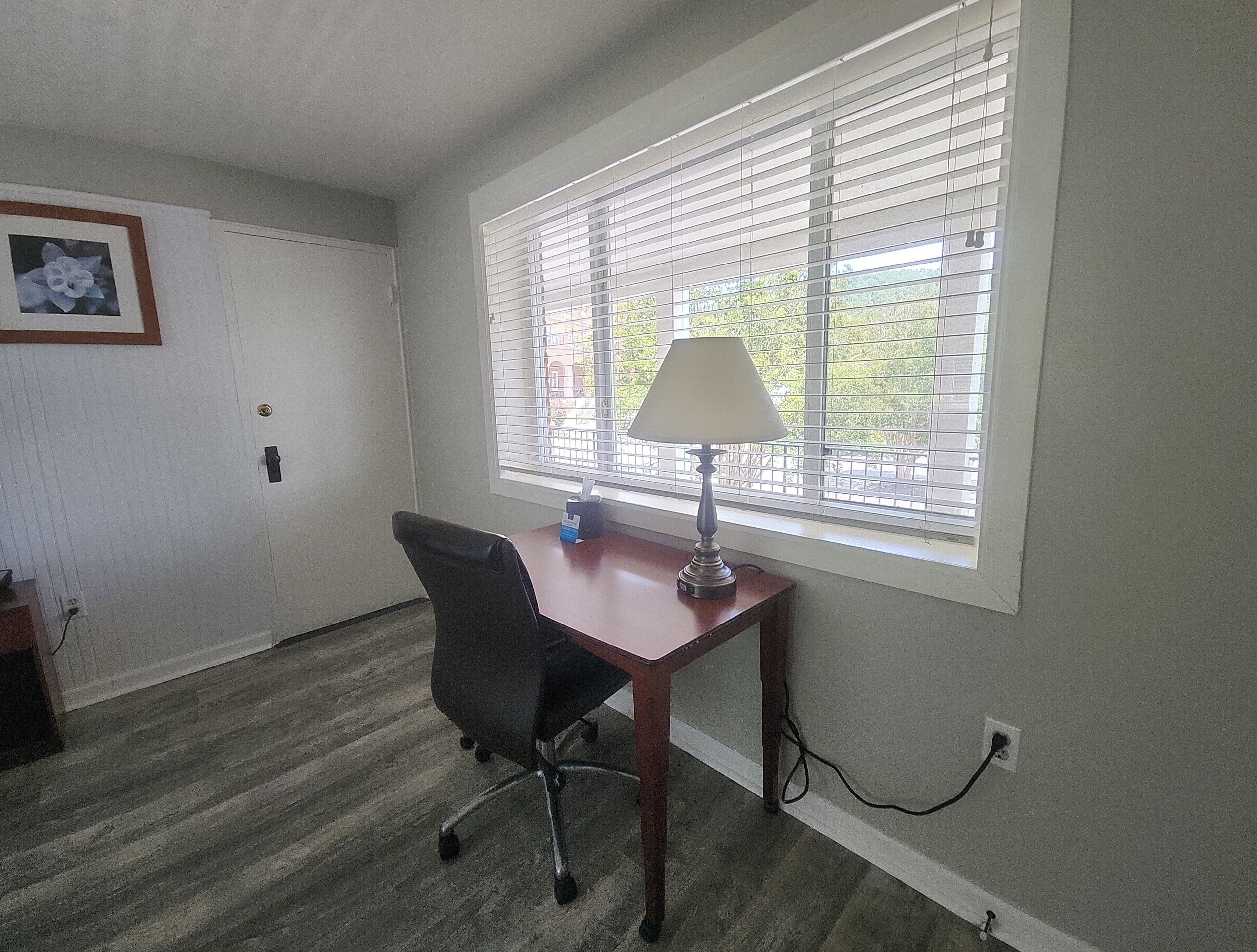 A room with a wooden desk, a black office chair, a window with blinds, and a wall-mounted picture.
