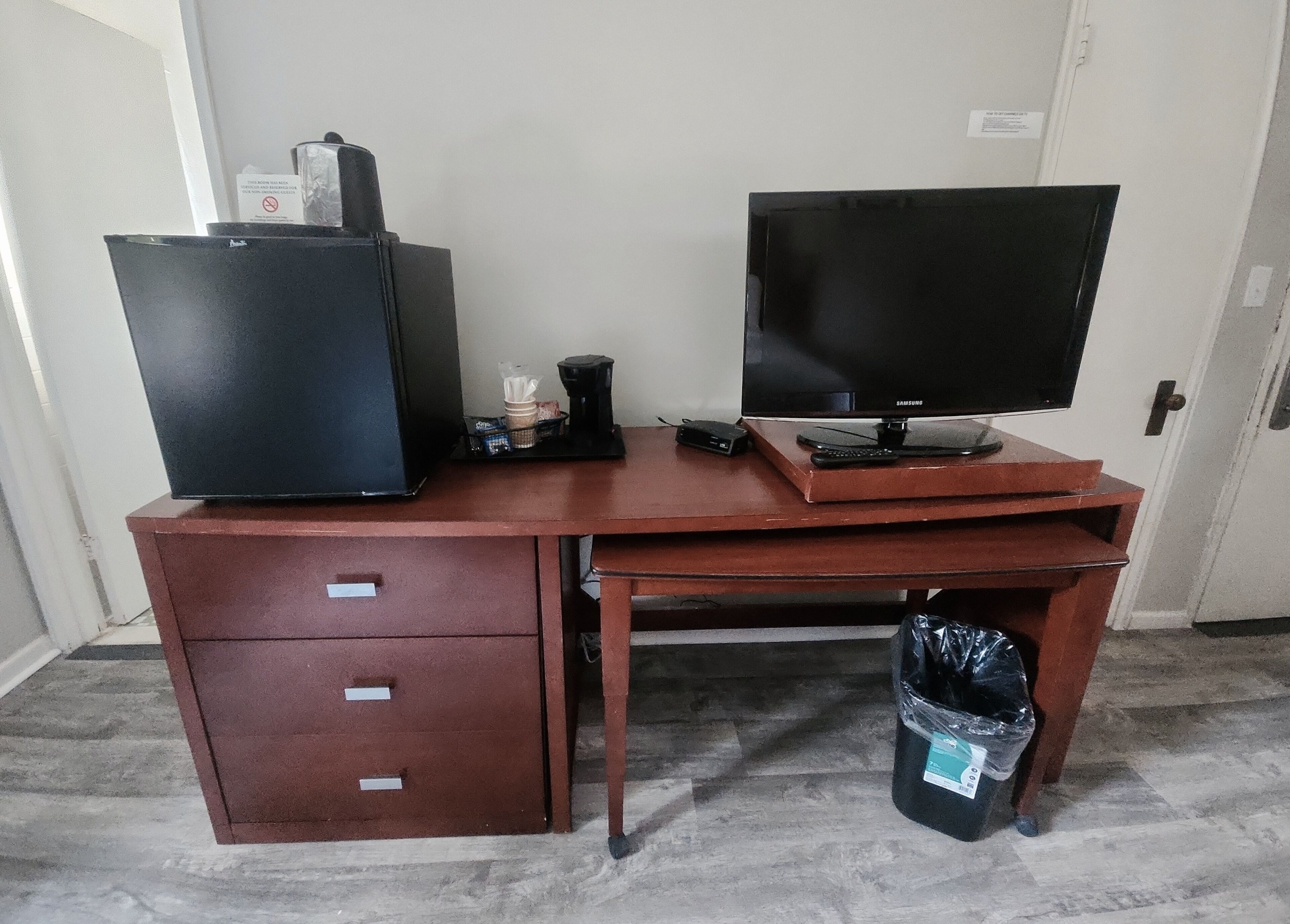 A wooden desk with a black refrigerator, a coffee maker, a remote control, and a trash bin.