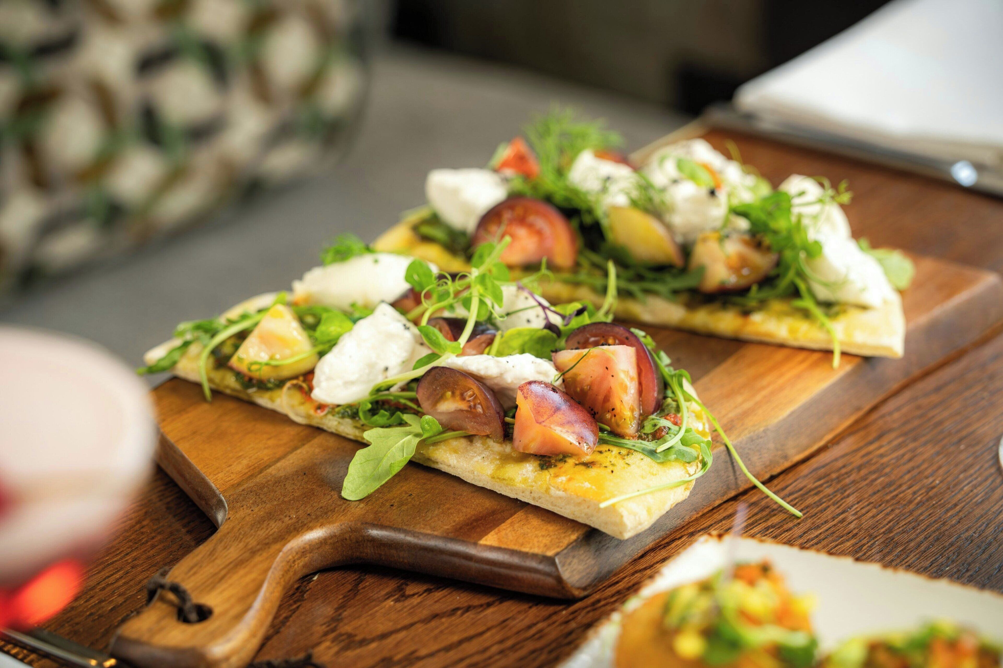 A wooden cutting board with a pizza topped with greens, tomatoes, and cheese.