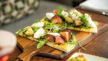 A wooden cutting board with a pizza topped with greens, tomatoes, and cheese.