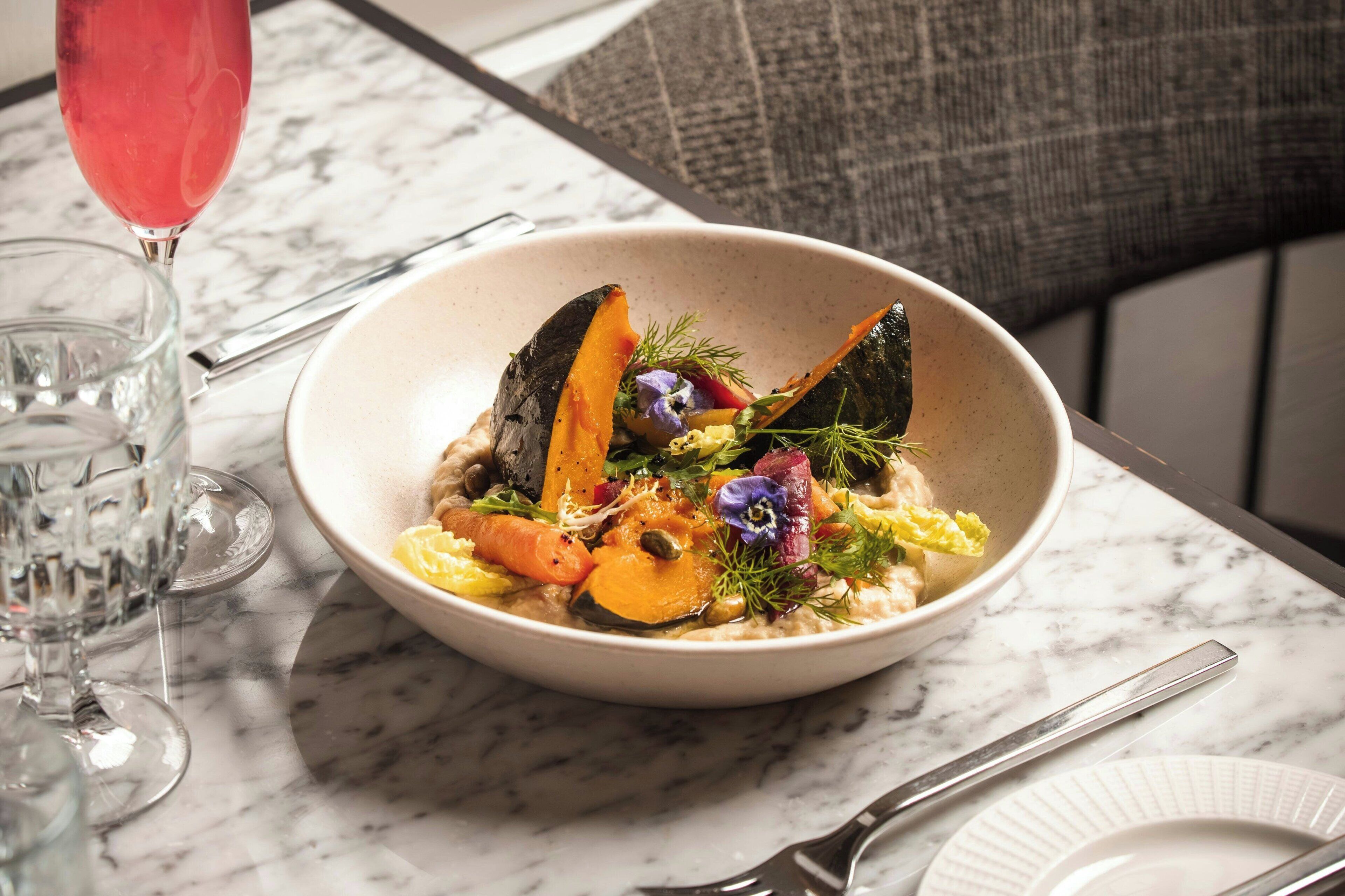 A bowl of food on a marble table with a glass of red drink and cutlery.