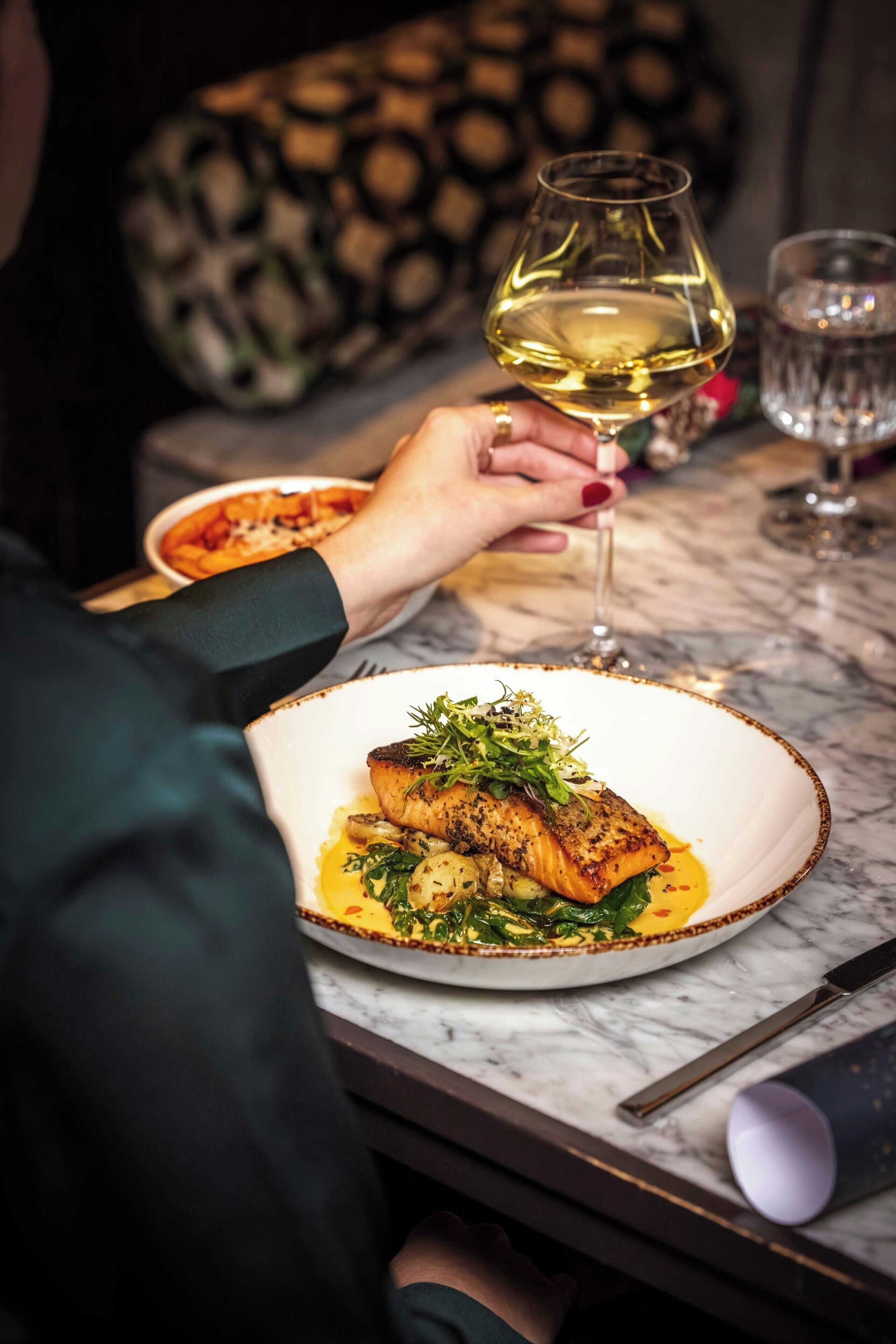 A person holding a wine glass, seated at a table with a plate of food.