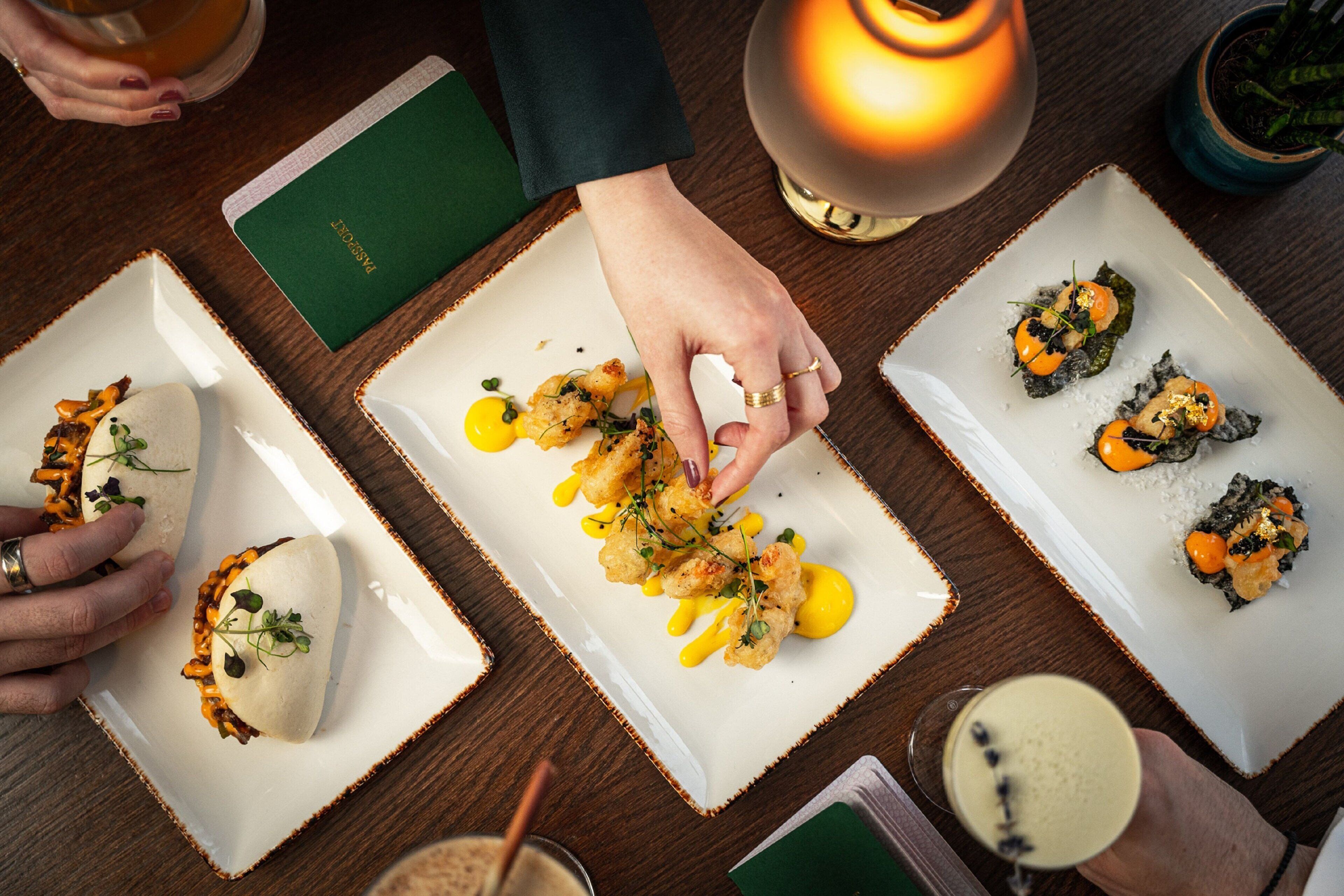 Una mesa de comedor con tres platos cuadrados de comida, una vela encendida y un menú.