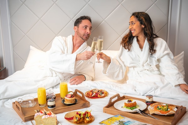 Un couple, dans une chambre d’hôtel, prend son petit-déjeuner : il y a des tartines, des viennoiseries et du jus de fruits.