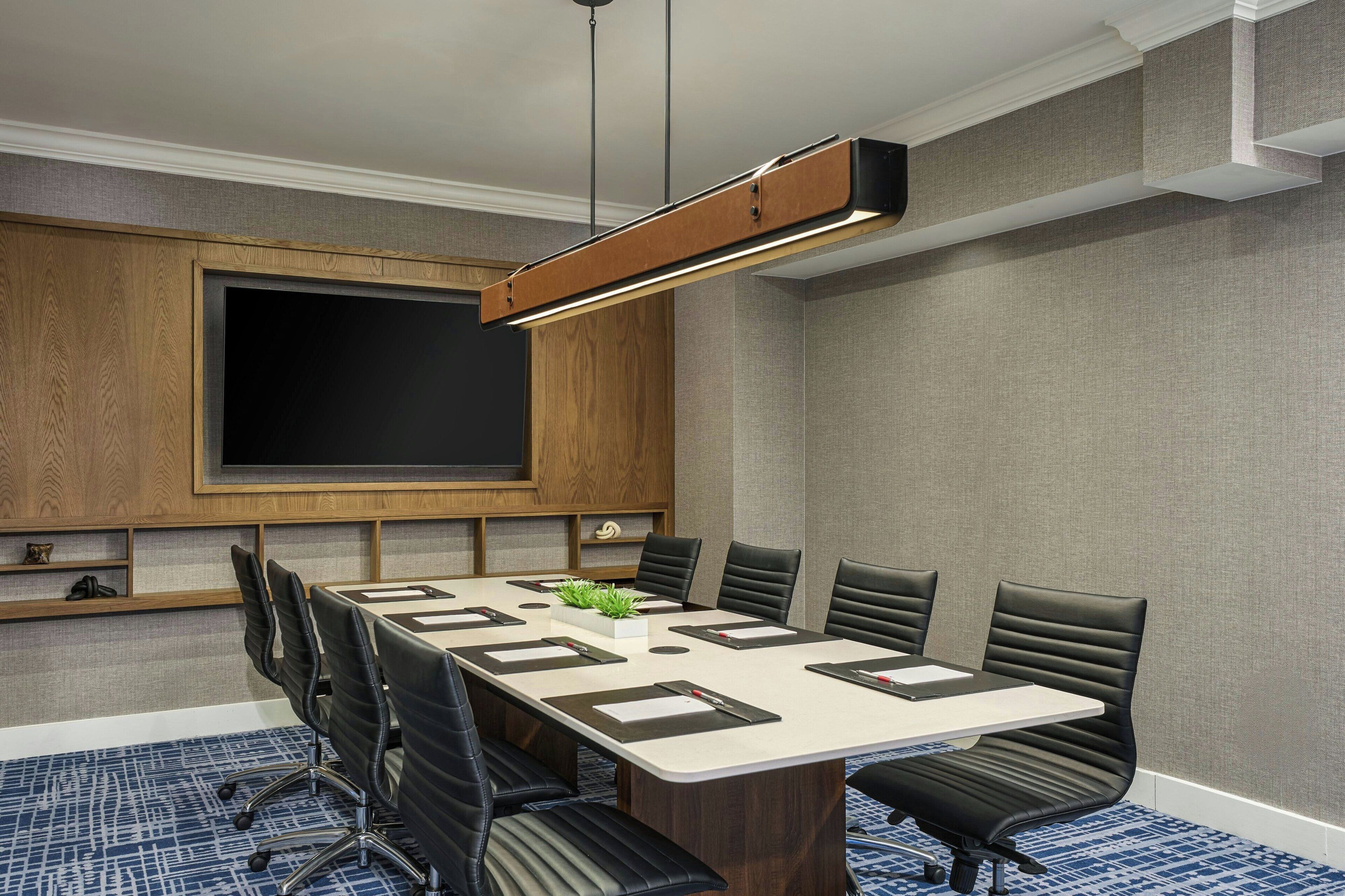 A conference room with a large table, black chairs, a projector screen, and a wooden panel wall.