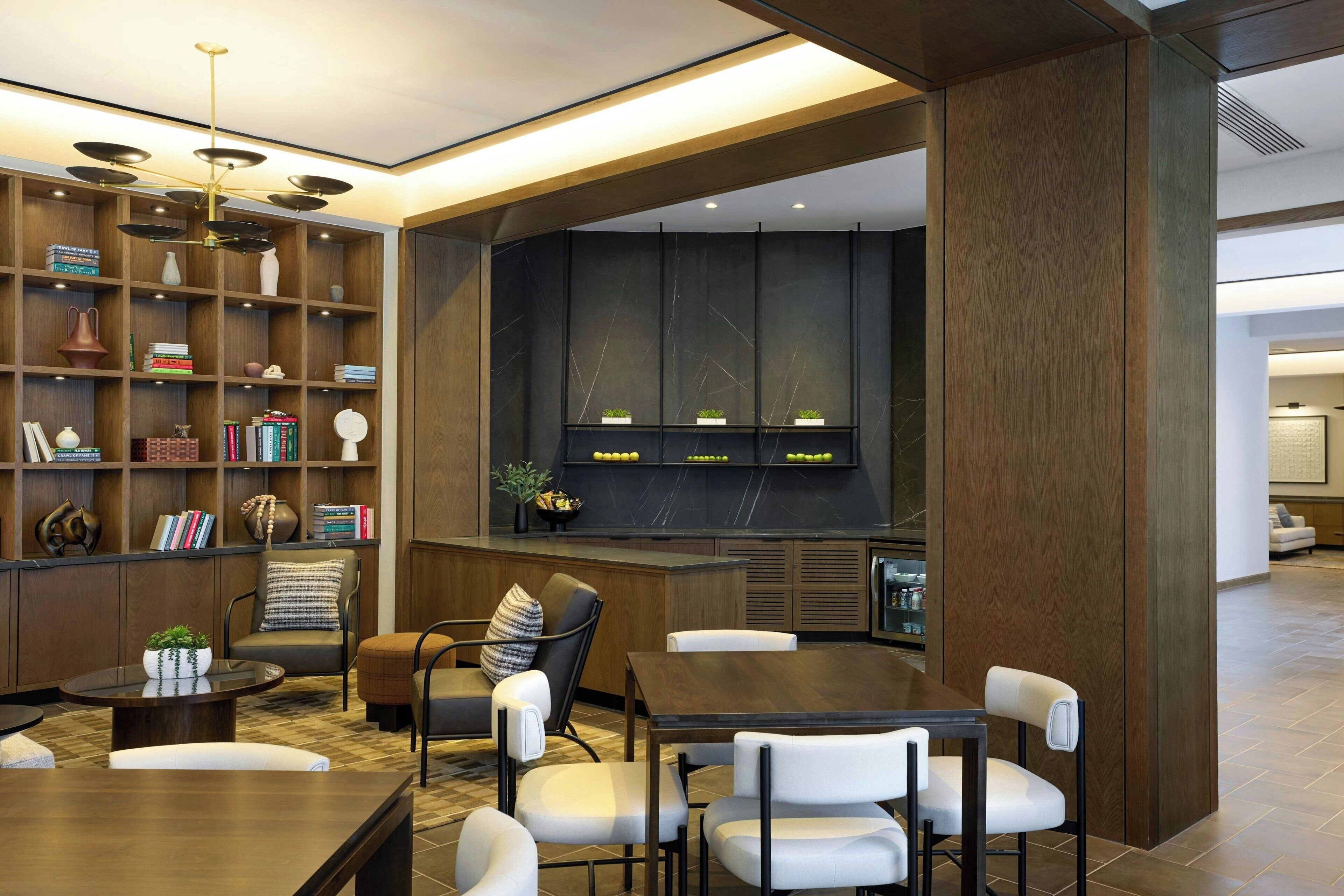 A modern interior with a wooden bookshelf, a dining area with white chairs, and a kitchen with a black backsplash.