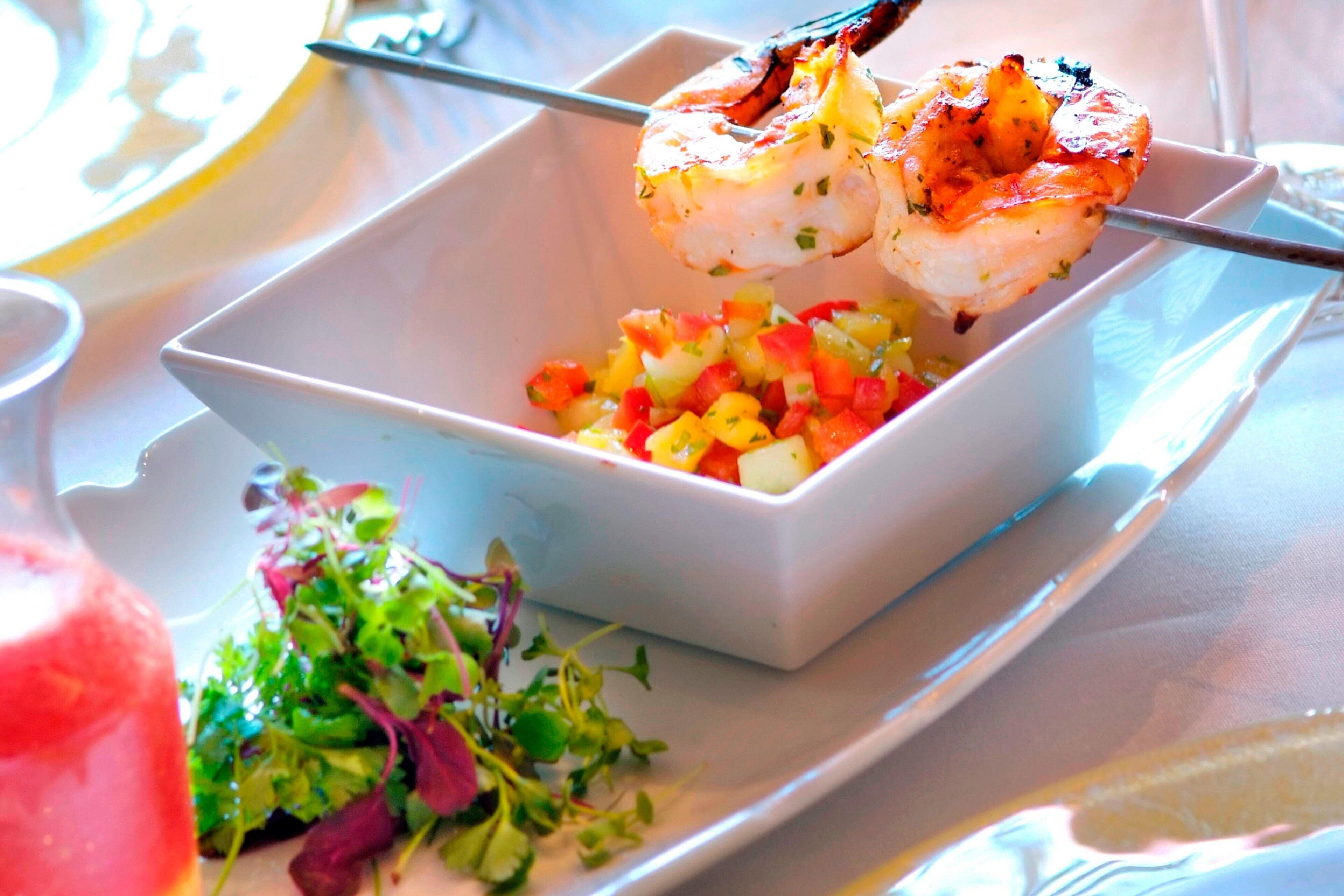 A plate with grilled shrimp skewers, a bowl of diced vegetables, and a side salad.