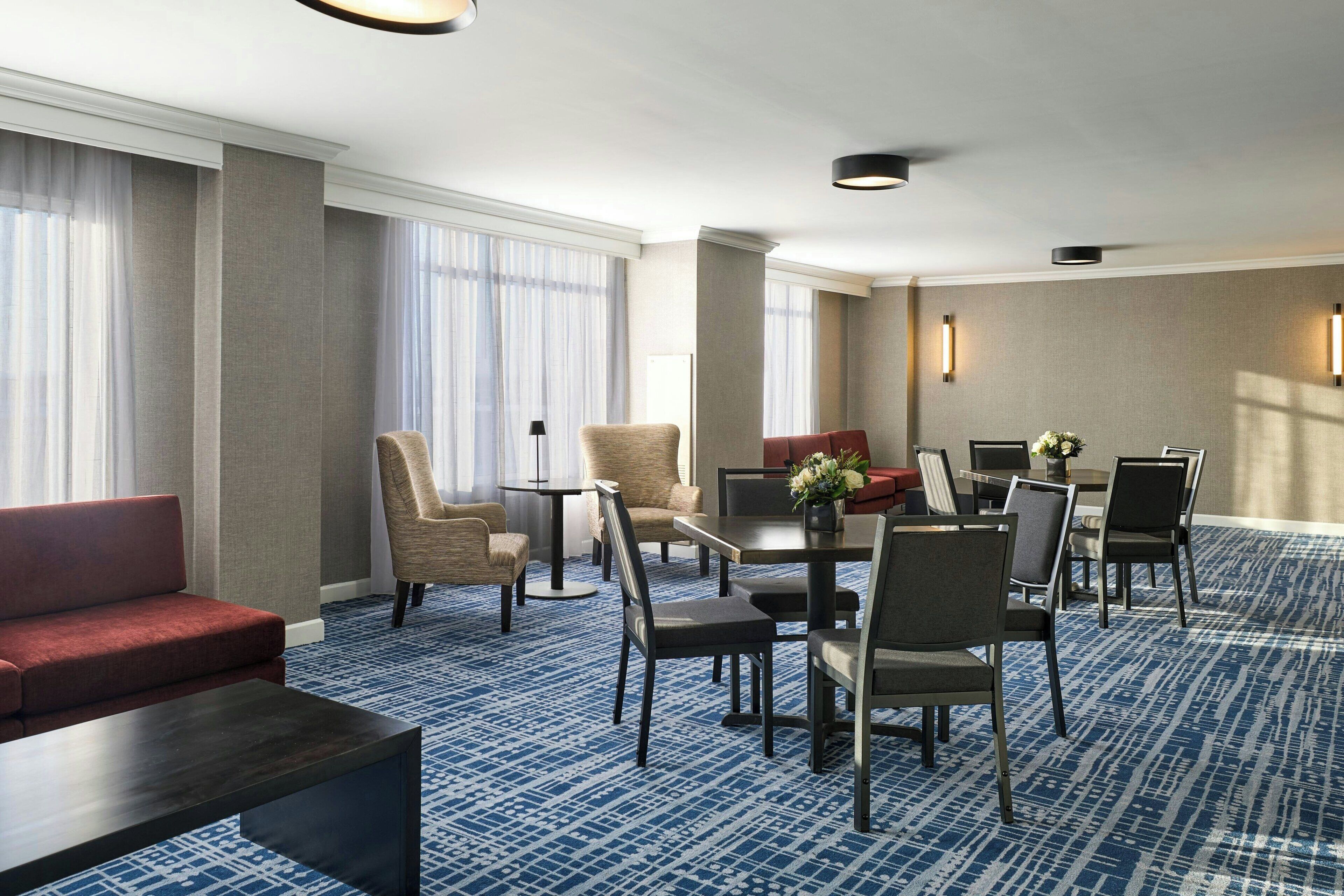 A hotel room with a red sofa, beige armchairs, a dining table with chairs, and a patterned carpet.