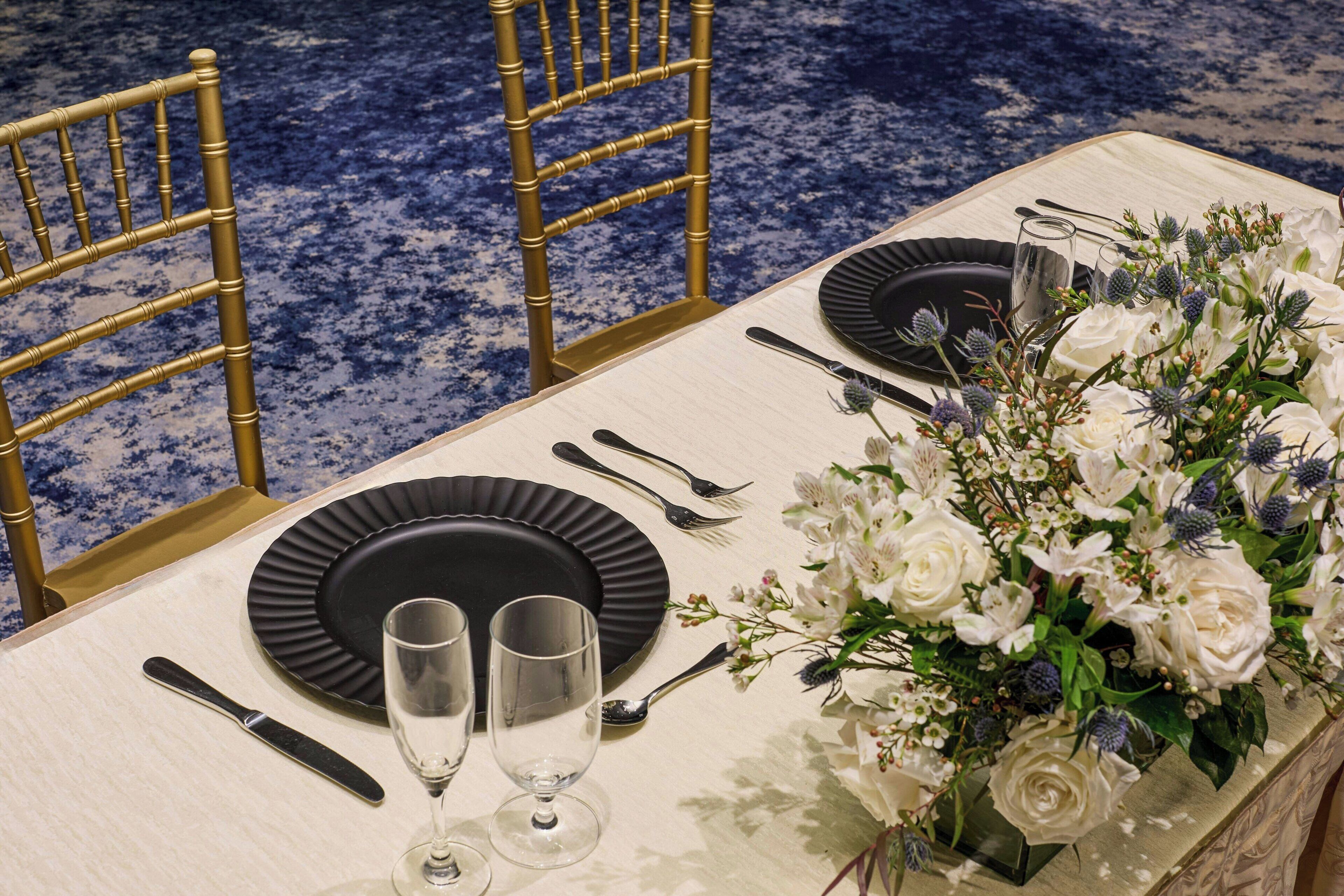 A table set with black plates, silverware, and two wine glasses, accompanied by a floral centerpiece.