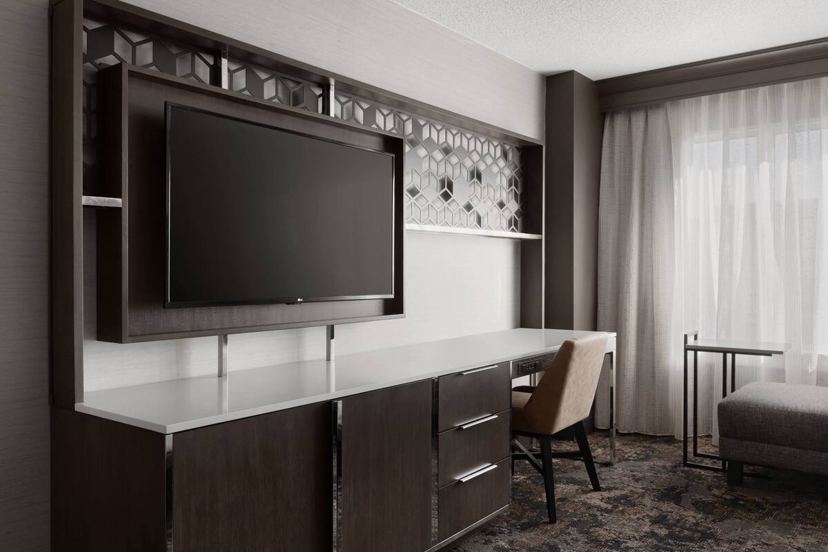 Una habitación de hotel moderna con un televisor de pantalla plana grande montado en la pared, un escritorio con una silla y una pequeña mesa con otra silla.