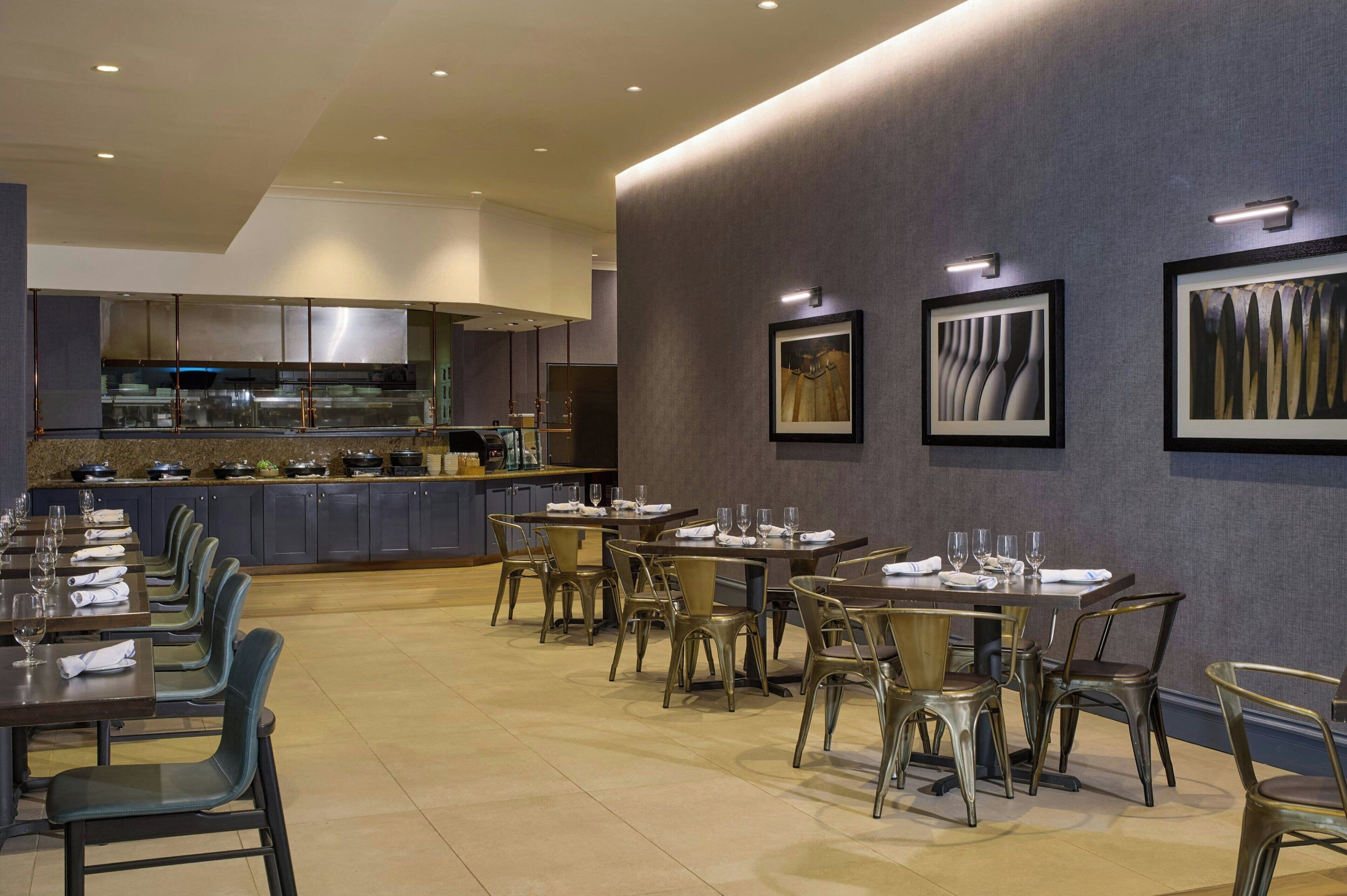 A modern dining area with multiple tables and chairs, a kitchen in the background, and framed artwork on the walls.
