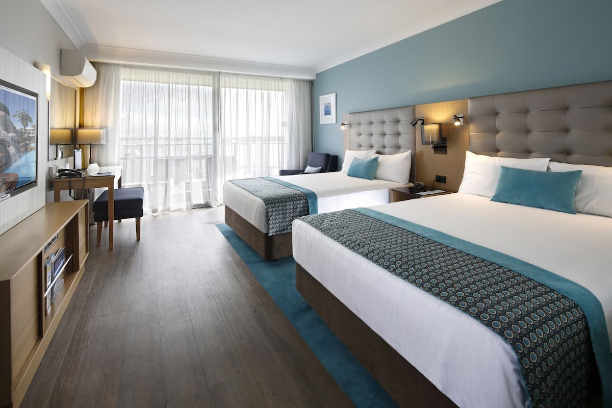 A hotel room with two beds, a desk, a chair, a TV, and a large window with curtains.